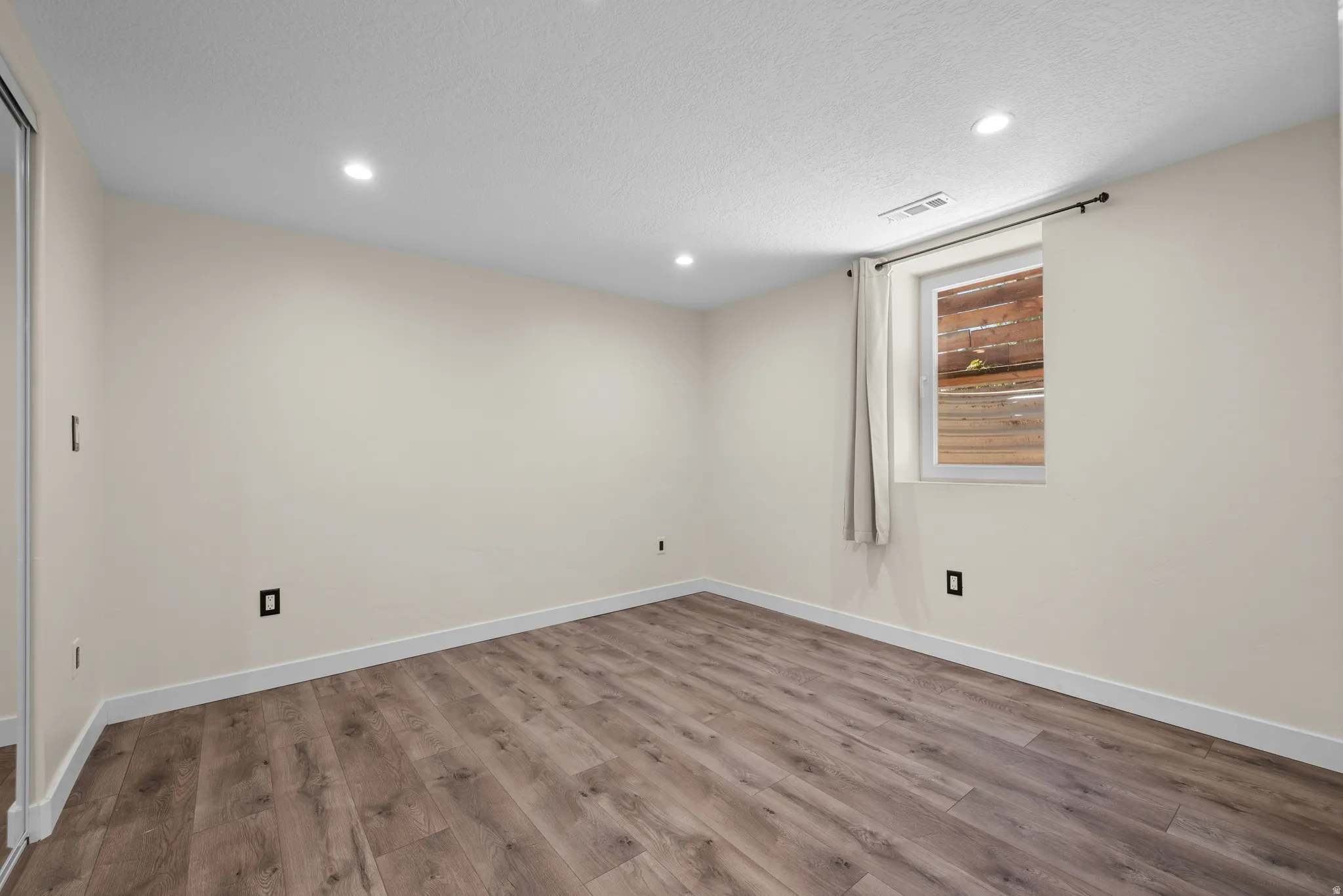 Spacious room featuring wood-finish flooring, recessed lighting, a window with a curtain rod, and a mirrored closet door