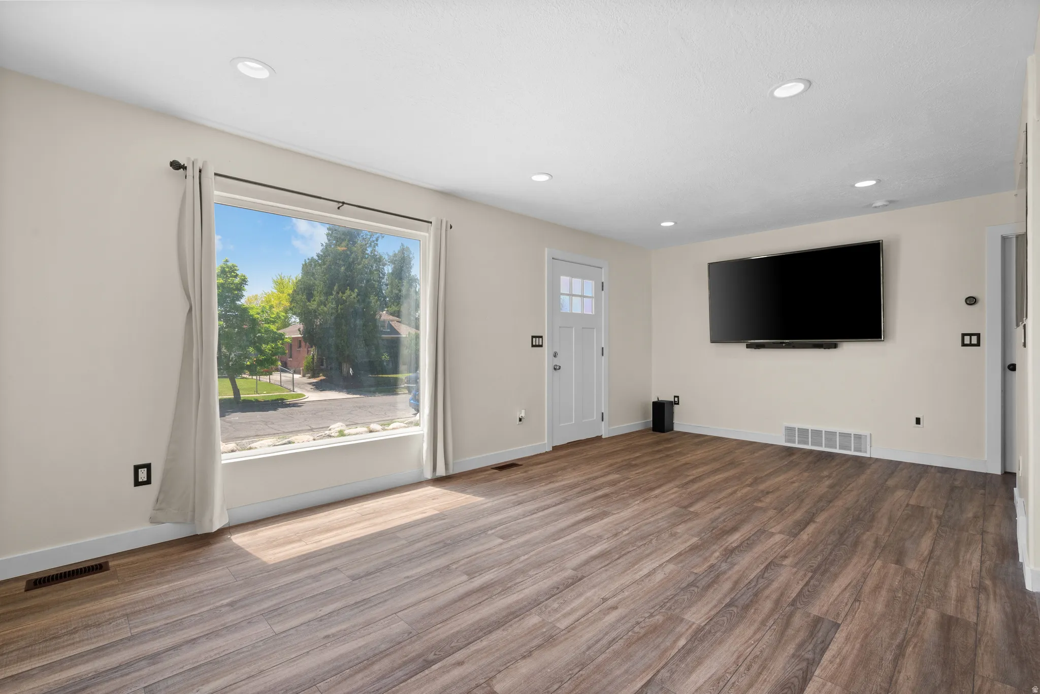 Spacious room featuring wood-finish flooring, a large picture window, recessed lighting, a white panel door with glass inserts, and light neutral wall tones