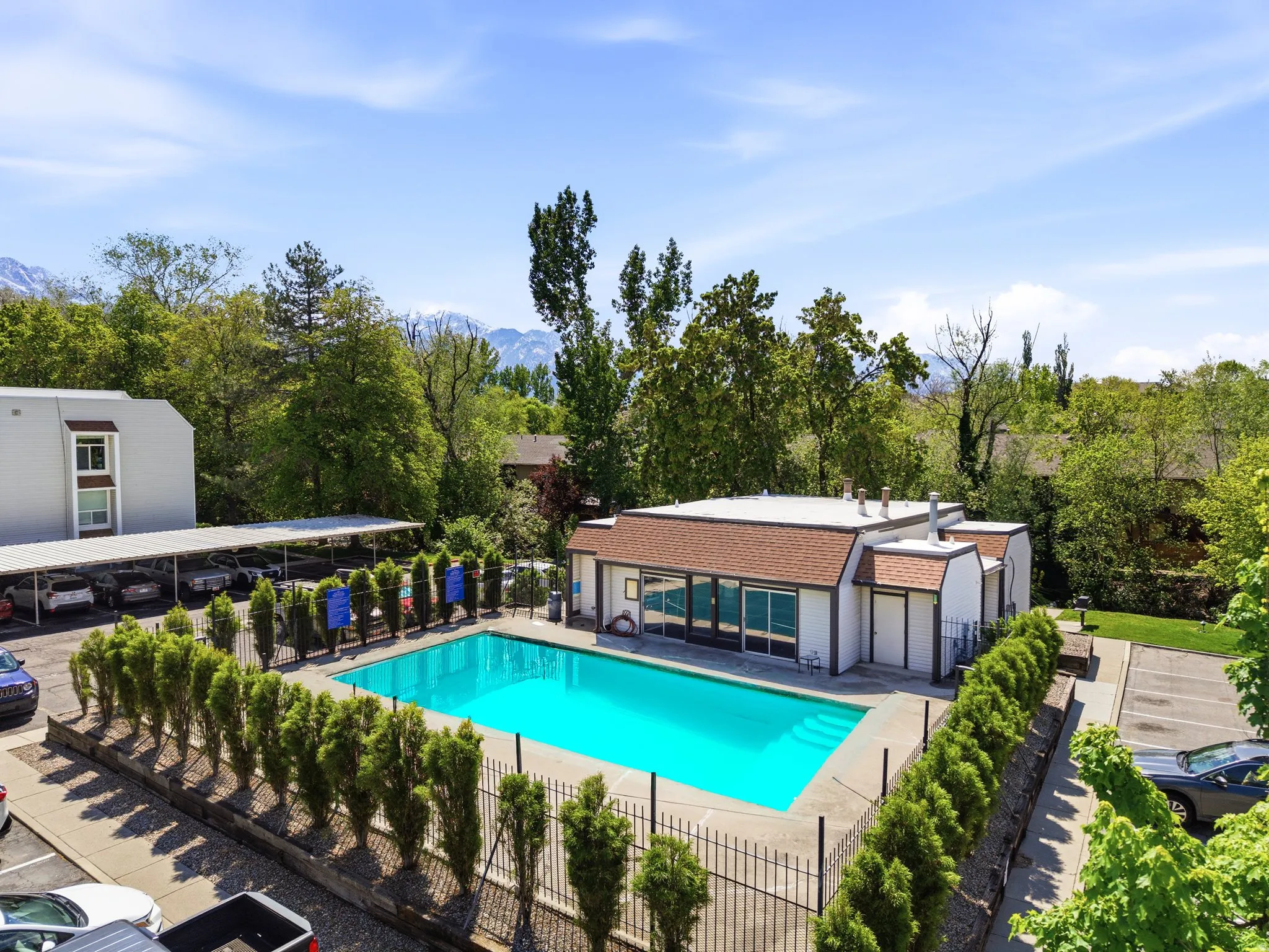 Fenced swimming pool area featuring a perimeter hedge, a pool house with a brown shingle roof, and surrounding paved parking