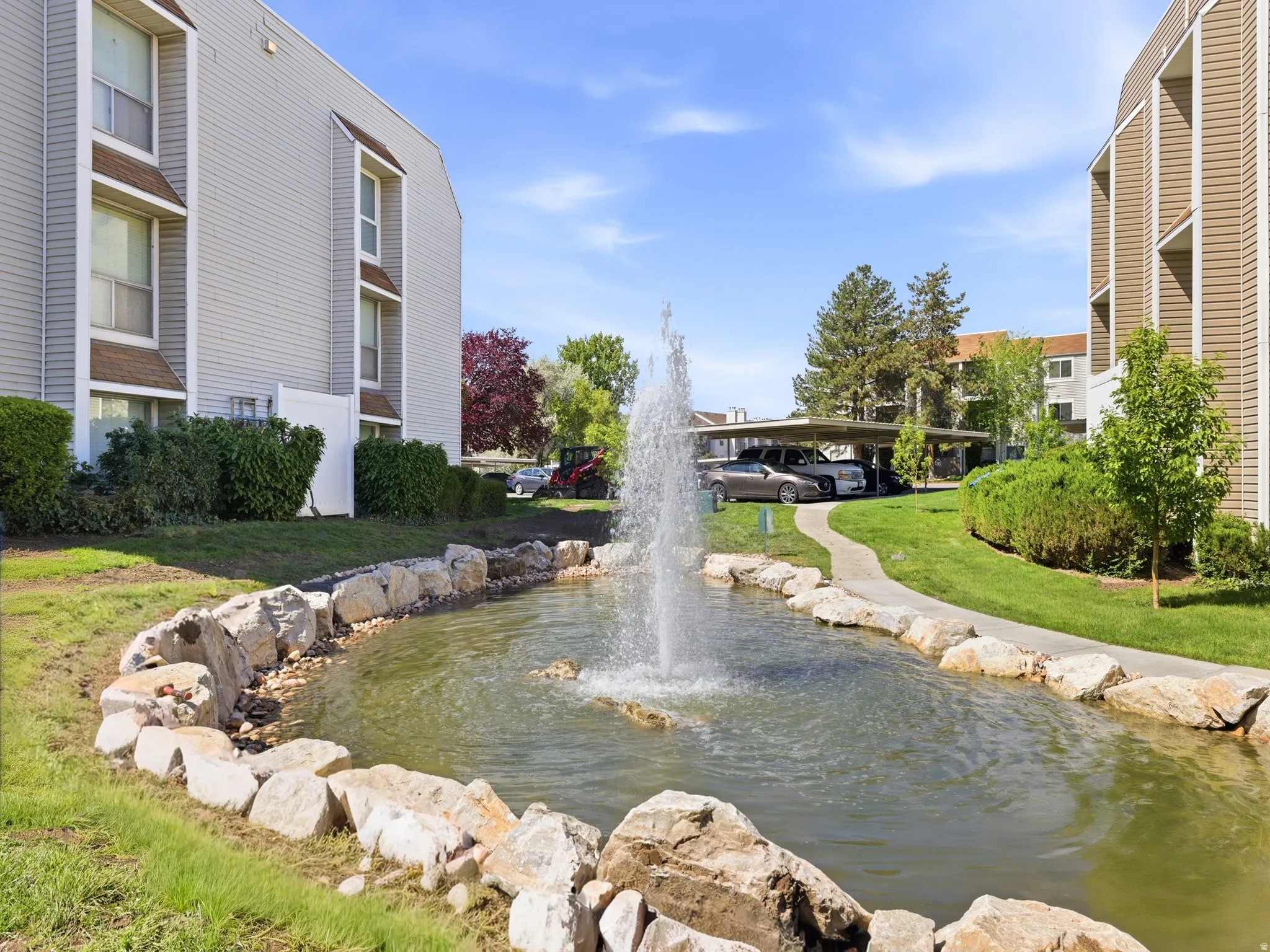 Manicured grounds featuring a rock-lined pond with an active fountain, a paved pathway, and established shrubs