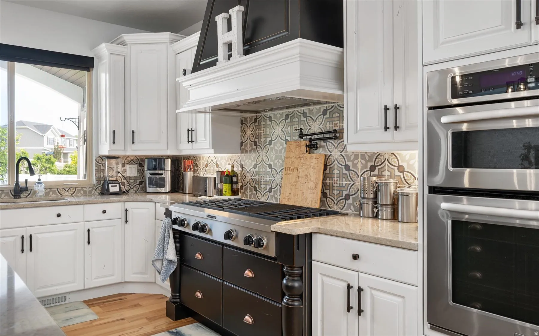 Gourmet kitchen featuring a professional-grade gas range, built-in double ovens, white cabinetry with black hardware, patterned tile backsplash, and light-toned countertops