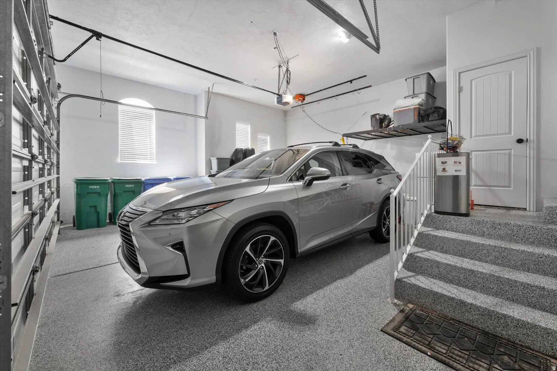 Spacious garage featuring a durable epoxy-coated floor, arched window with blinds, overhead door, built-in shelving, and direct entry to interior stairs with a white railing