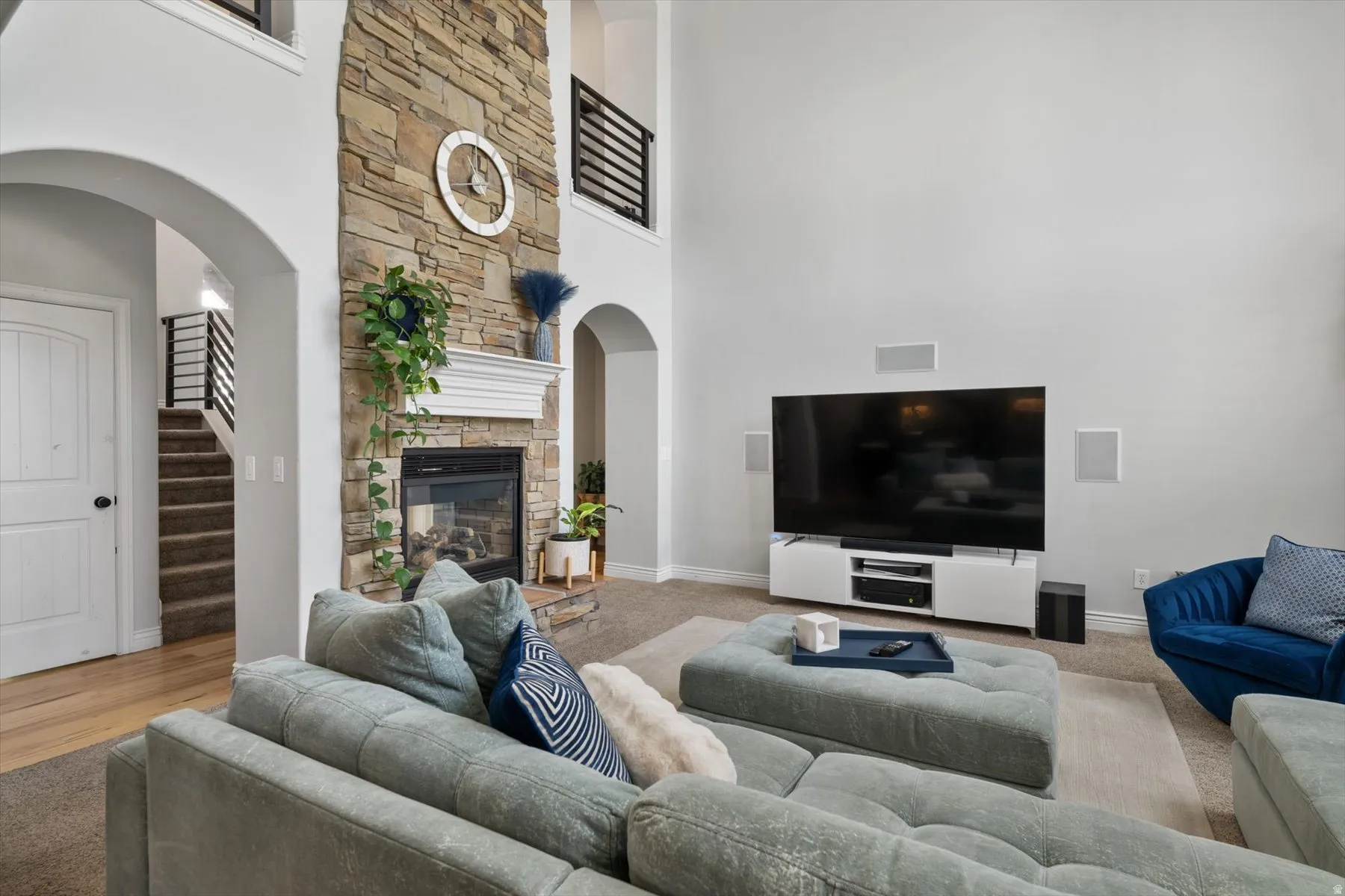 Two-story living area featuring a stacked stone fireplace with a white mantel, arched doorways, a carpeted staircase with dark metal railings, light wood-finish flooring, and built-in wall speakers