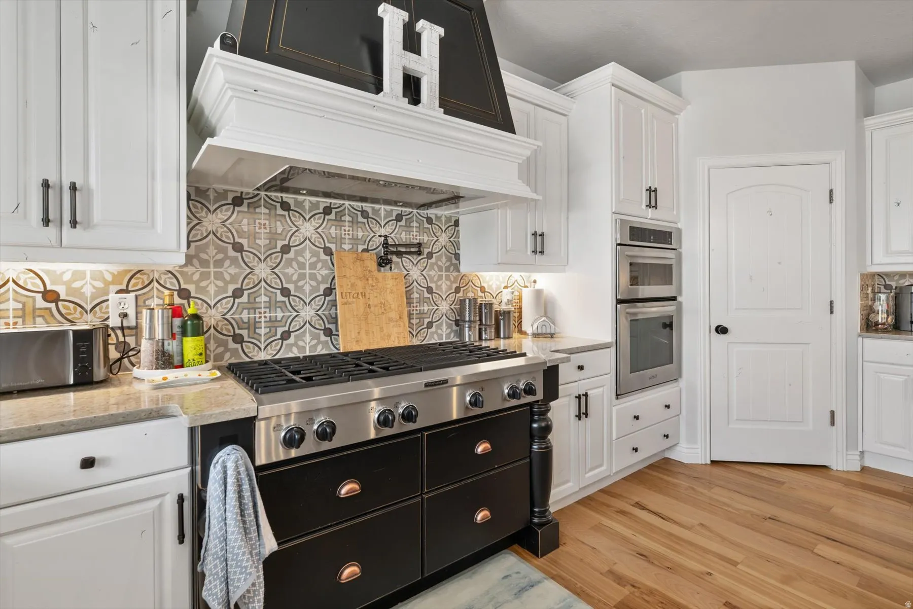 Gourmet kitchen featuring a professional-grade range, custom black and white cabinetry, patterned tile backsplash, built-in double ovens, and wood-finish flooring