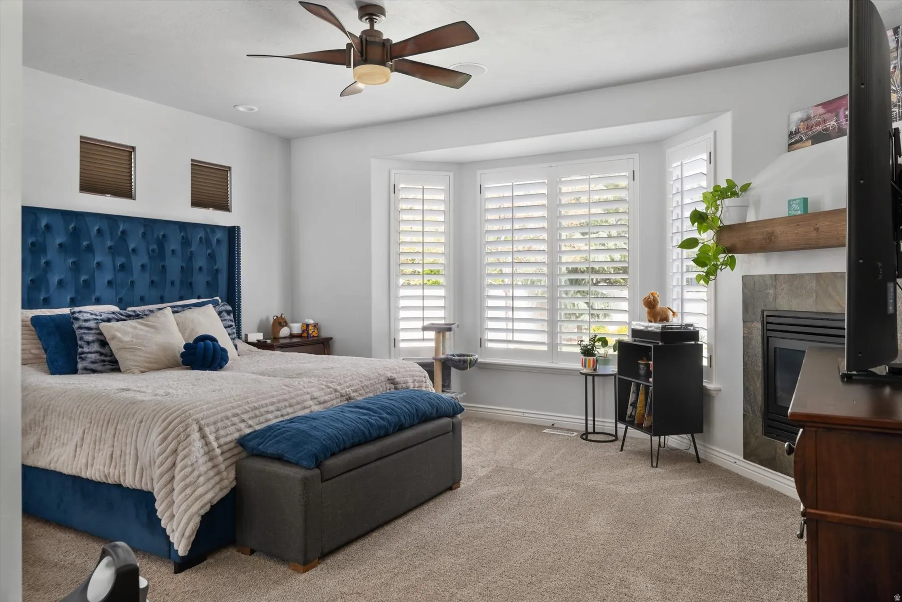 Primary Bedroom with recessed lighting and a ceiling fan fixture illuminate a spacious room featuring bay windows with white plantation shutters, a tiled fireplace with a wood mantle, and light-toned carpeting