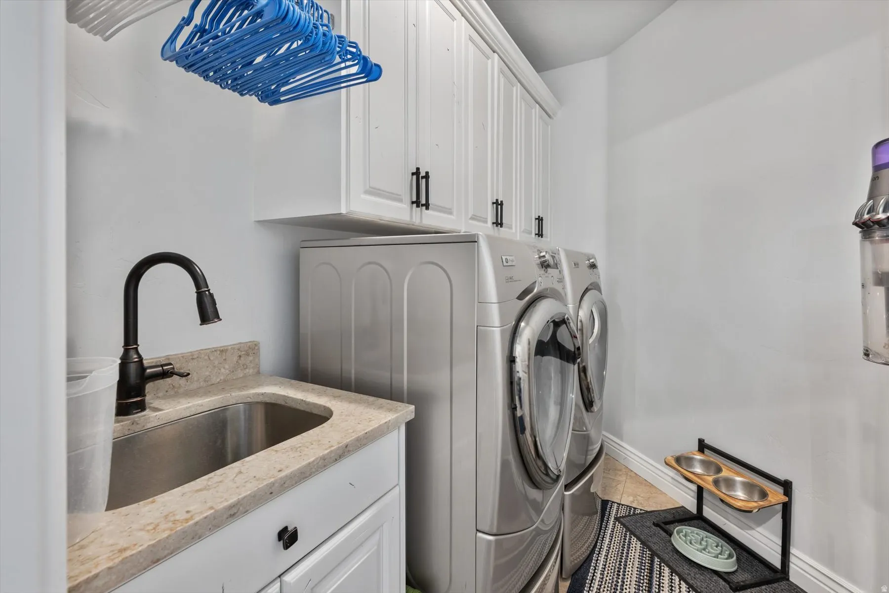 Laundry area featuring a utility sink, a dark bronze gooseneck faucet, light-toned stone-finish countertop, white cabinetry with dark hardware, and tile flooring