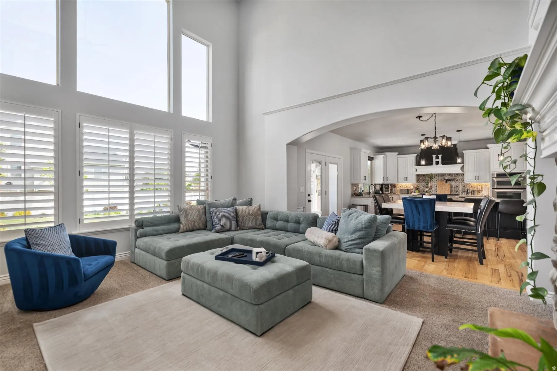 Spacious living area featuring high ceilings, tall windows with plantation shutters, and an arched doorway opening to a kitchen with wood-finish flooring