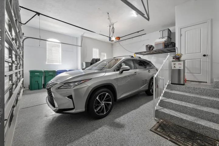 Spacious garage featuring a durable epoxy-coated floor, arched window with blinds, overhead door, built-in shelving, and direct entry to interior stairs with a white railing