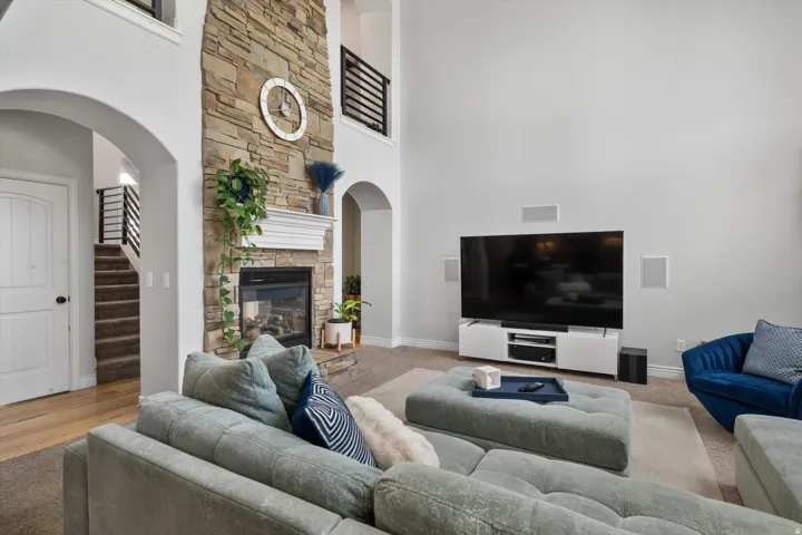 Two-story living area featuring a stacked stone fireplace with a white mantel, arched doorways, a carpeted staircase with dark metal railings, light wood-finish flooring, and built-in wall speakers
