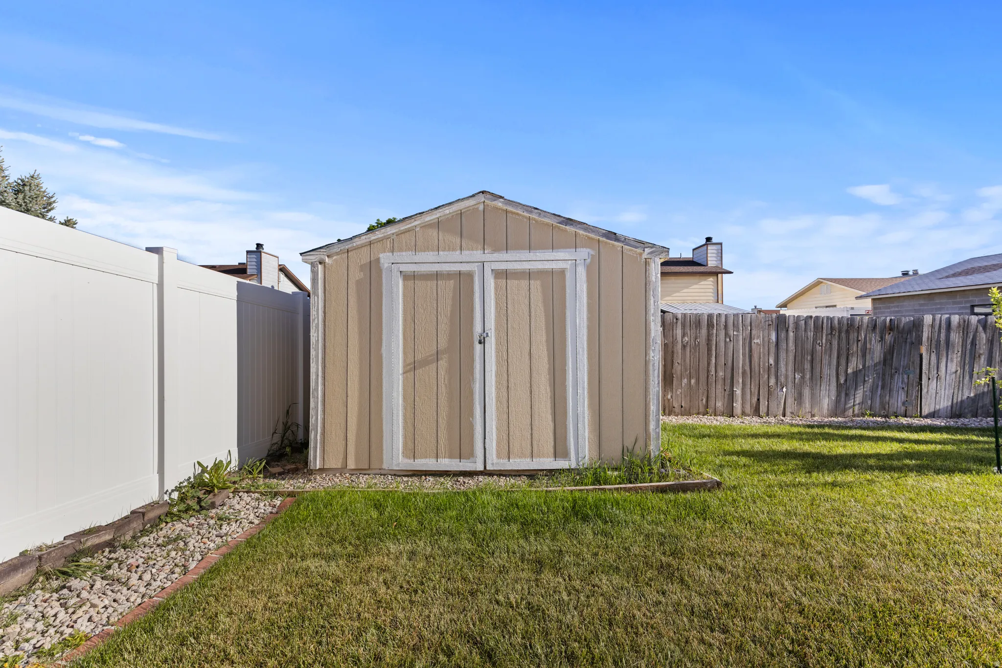 Backyard featuring a tan storage shed with double doors, a white privacy fence, a wood picket fence, a manicured lawn, and a decorative rock border