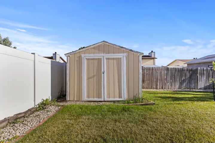 Backyard featuring a tan storage shed with double doors, a white privacy fence, a wood picket fence, a manicured lawn, and a decorative rock border