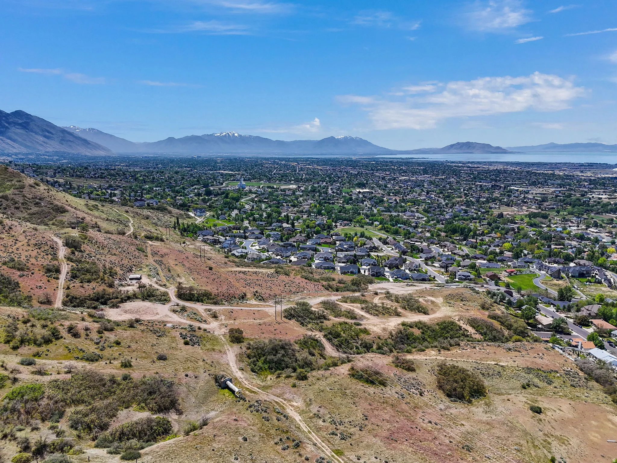 Expansive hillside property featuring undeveloped terrain with distant residential areas, mountains, and a large body of water under a clear blue sky