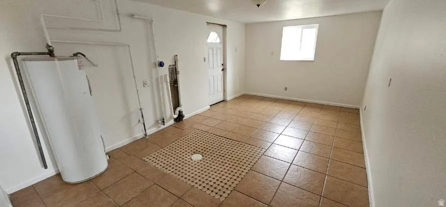 Utility space featuring a water heater, visible plumbing, a tiled floor with a central drain, a white paneled door, and a single window