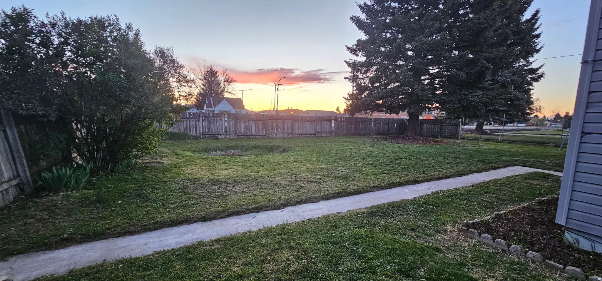 Expansive lawn area featuring a concrete pathway, mature trees, and a perimeter fence