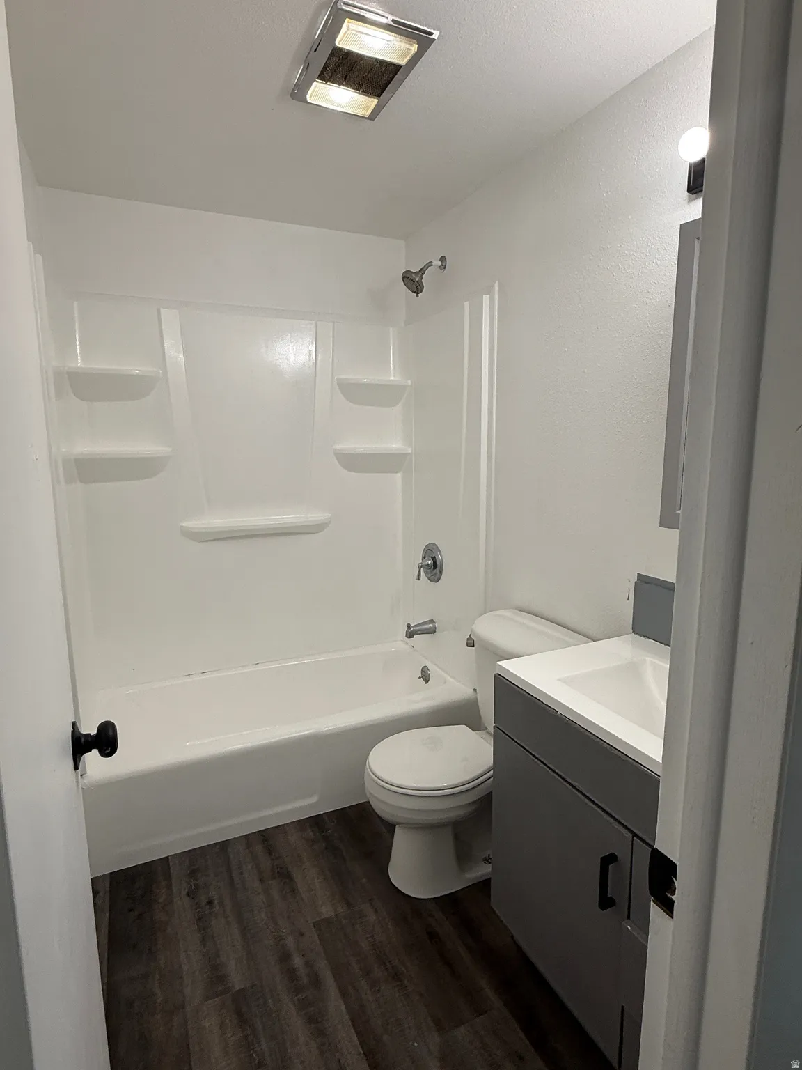 Bathroom featuring a white bathtub and shower surround, a toilet, a gray vanity with an integrated sink, and wood-finish flooring