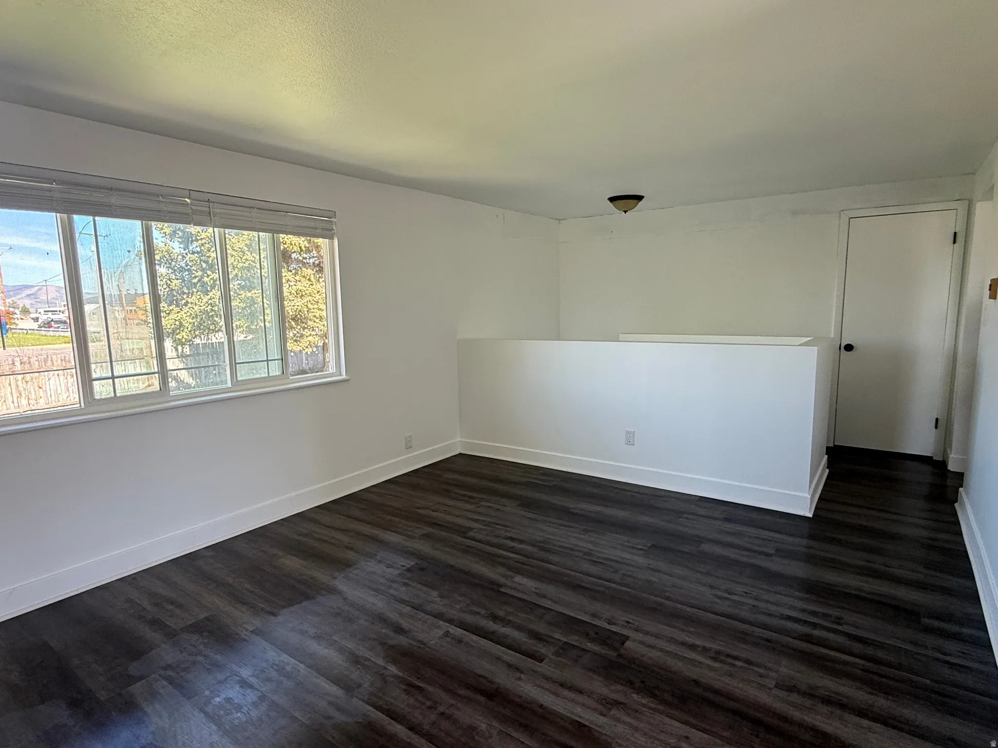Spacious room featuring dark wood-finish flooring, white baseboards, and a large multi-pane window with blinds