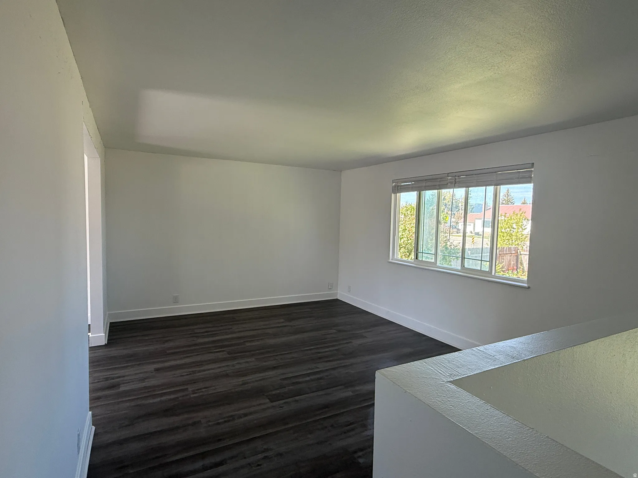 Spacious room with dark wood-finish flooring, white walls, and a large window with white blinds