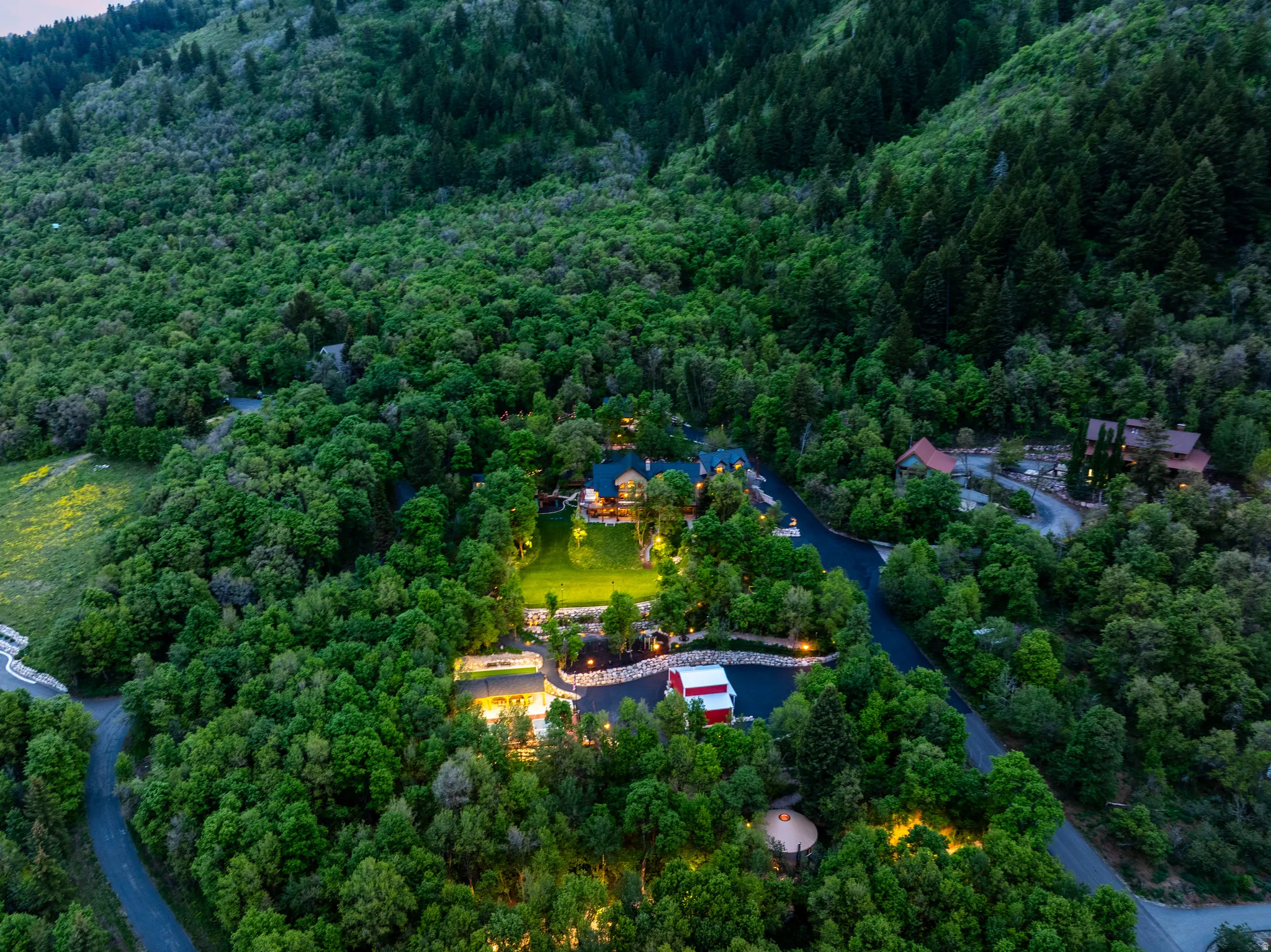 Expansive property nestled among mature trees, featuring multiple structures, paved driveways, a central illuminated lawn, and surrounding forested hills