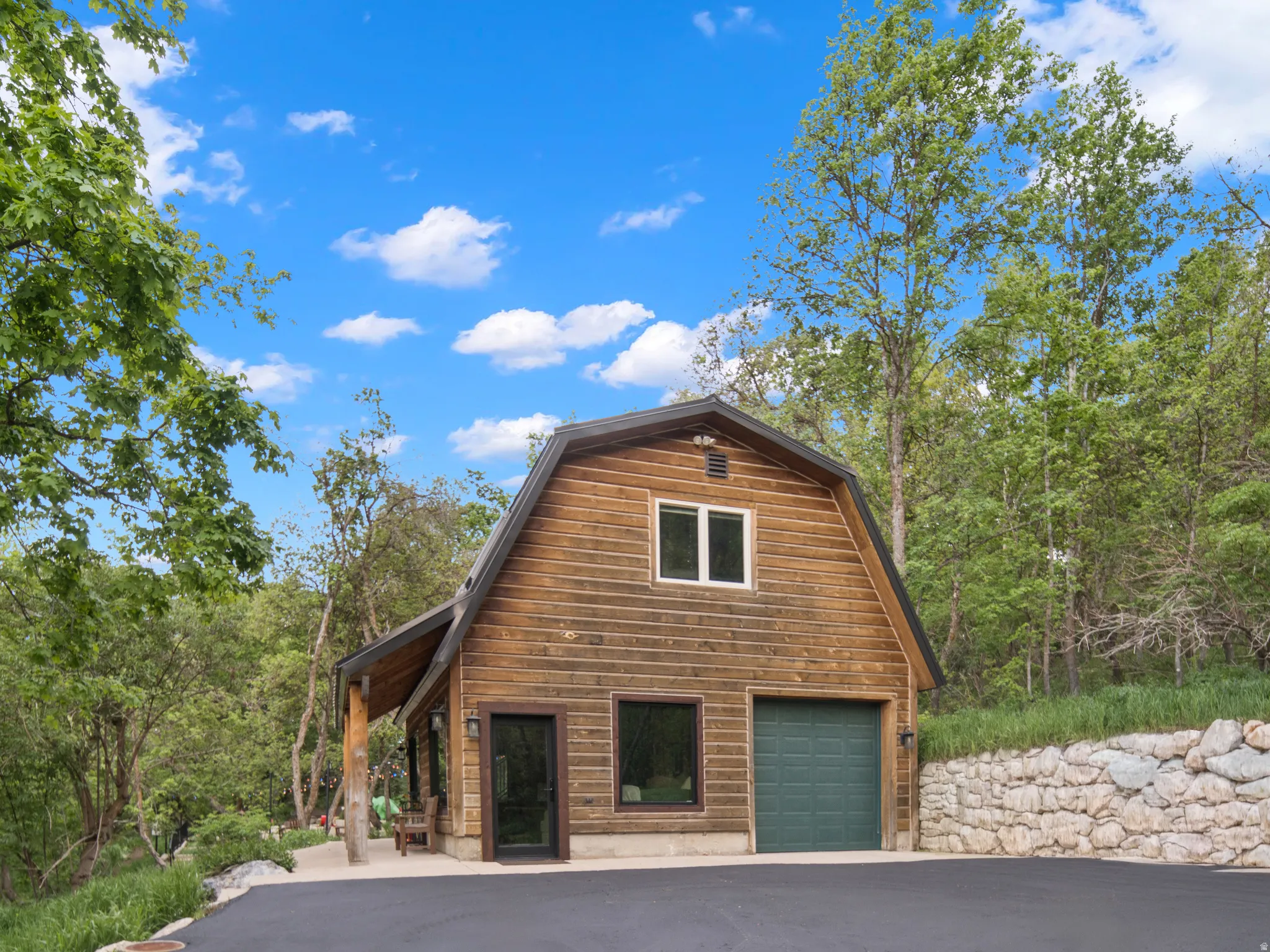 Wood-clad gambrel-style structure featuring a green garage door, covered porch, and upper-level window