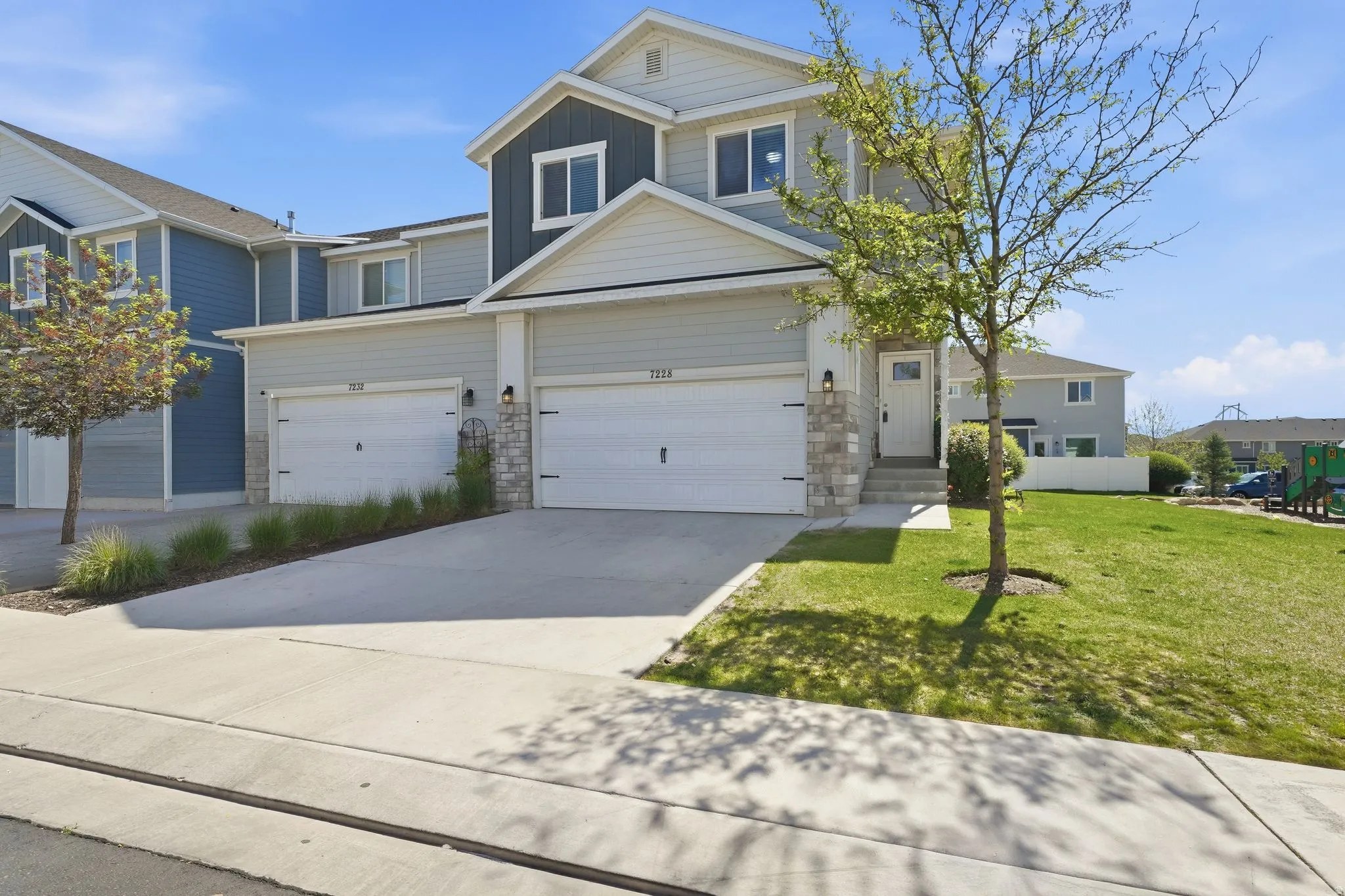 Contemporary exterior featuring light grey siding with dark grey accents, stone veneer detailing, a two-car garage, manicured lawn, and a light-colored entry door with window detail