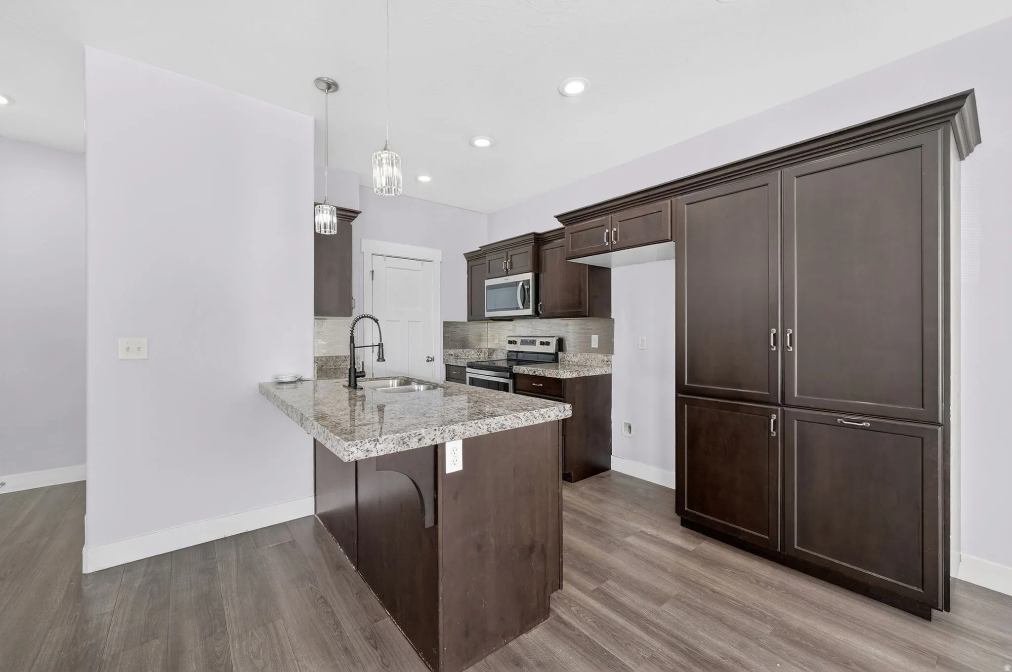 Kitchen featuring dark wood cabinetry, a central island with a granite-style countertop, stainless steel appliances, pendant lighting, and wood-finish flooring