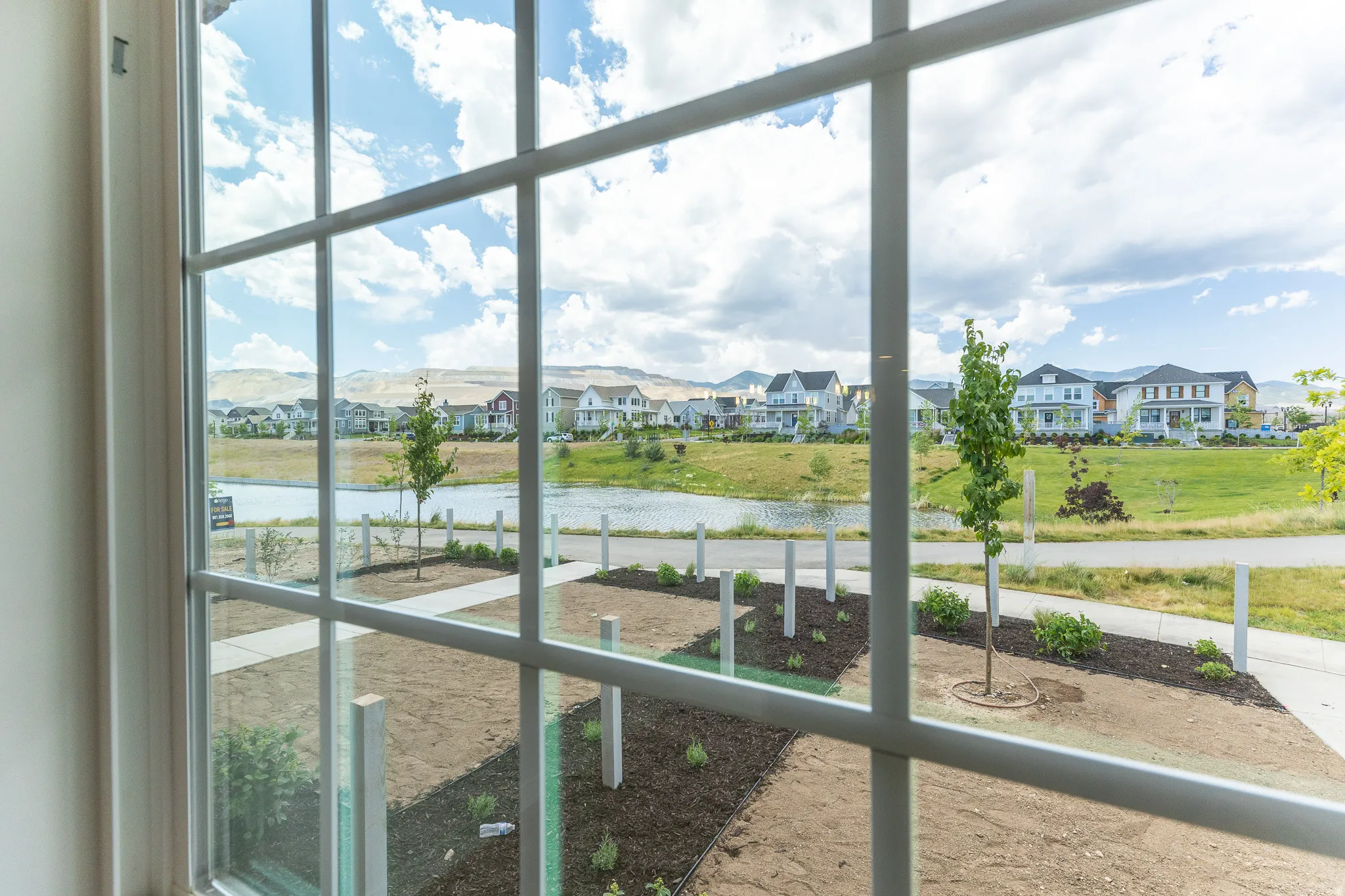 Gridded window overlooking a landscaped area with a paved path, a body of water, and residential structures in the distance