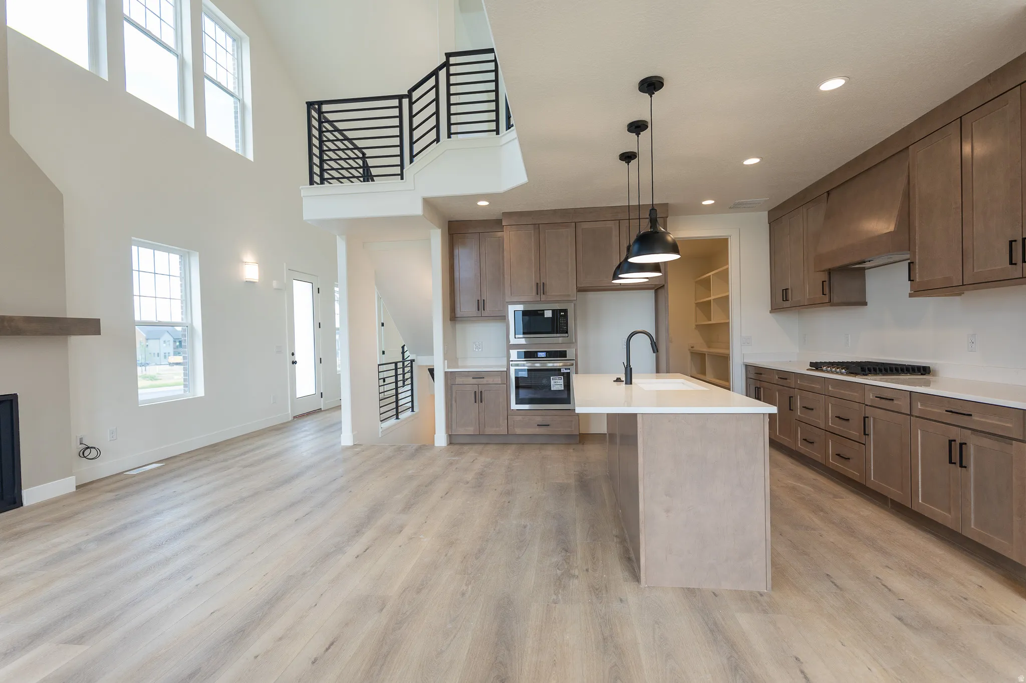 Open-concept interior featuring light wood-finish flooring, a two-story ceiling with high windows, a kitchen island with pendant lighting, light wood cabinetry, and a black metal staircase railing