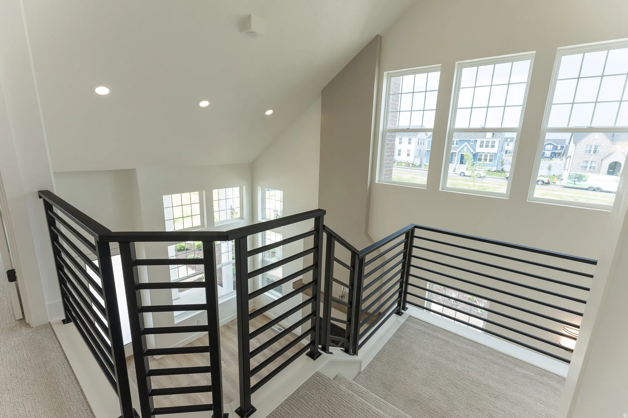 Open concept upper landing featuring black horizontal bar railings, three large grid-pattern windows, vaulted ceilings with recessed lighting, and neutral carpeting