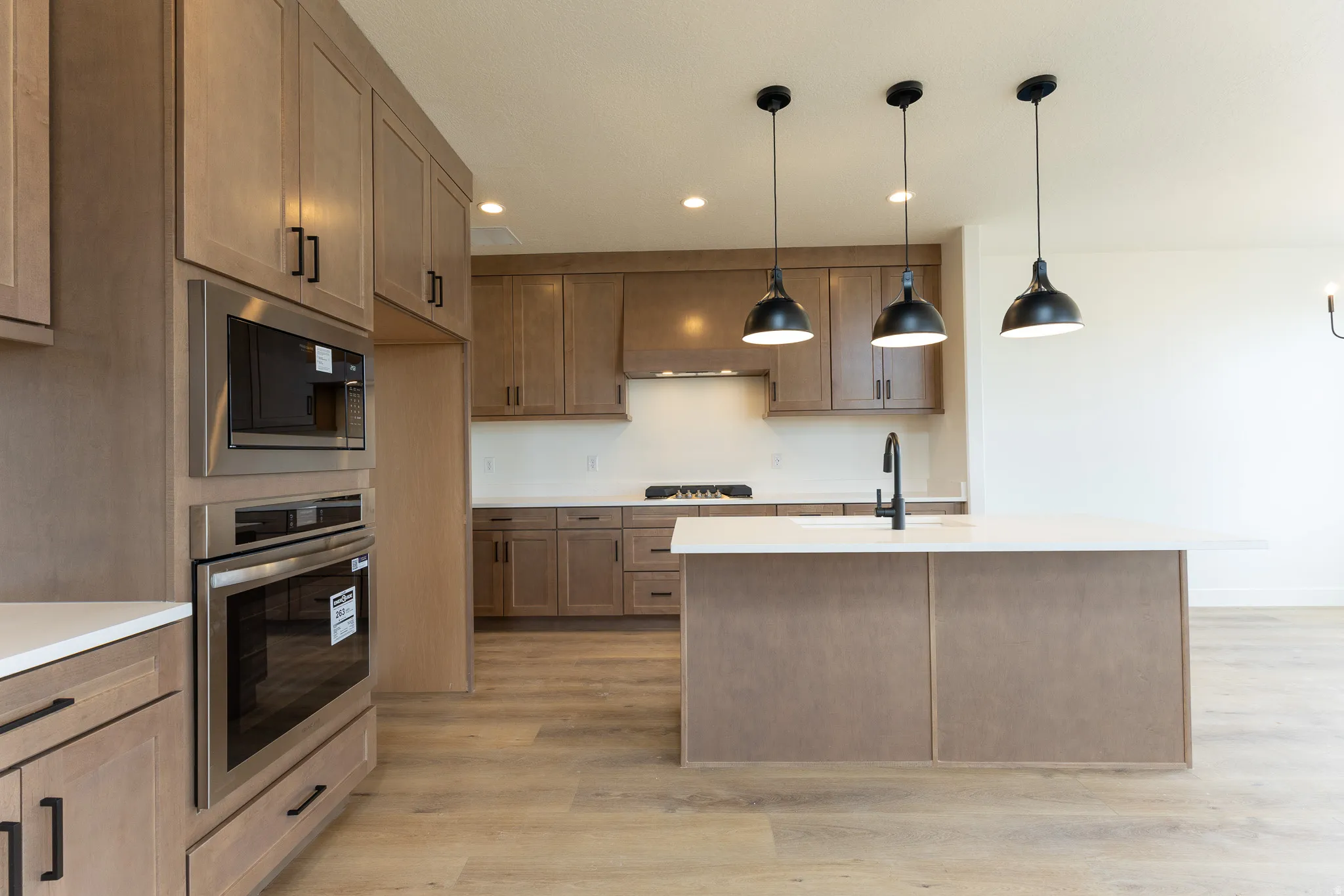 Modern kitchen featuring wood-finish flooring, light wood cabinetry, stainless steel built-in appliances, a central island with a sink, and pendant lighting fixtures