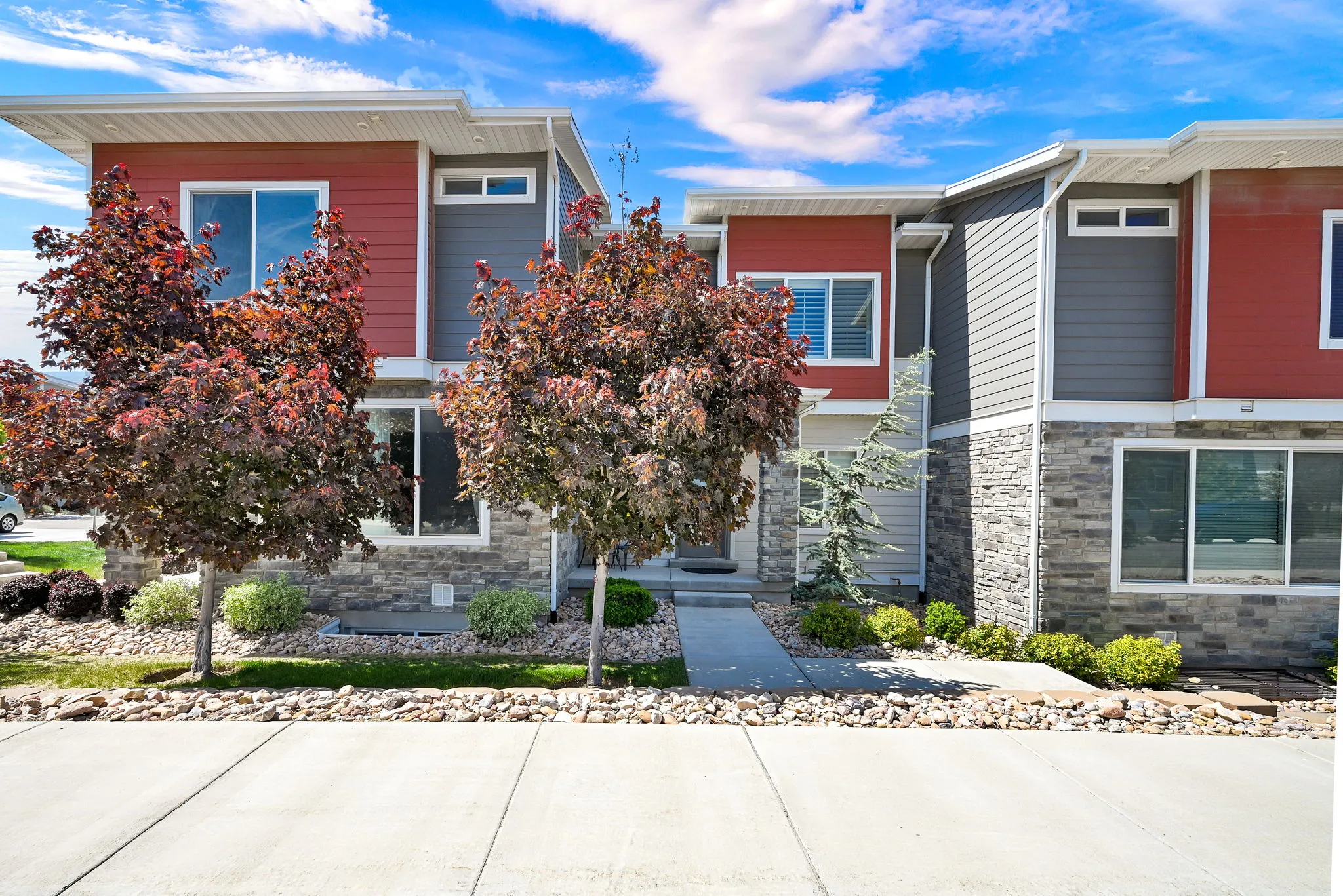 Modern multi-level exterior featuring red and gray siding, stone veneer accents, large windows, mature landscaping, and a concrete walkway