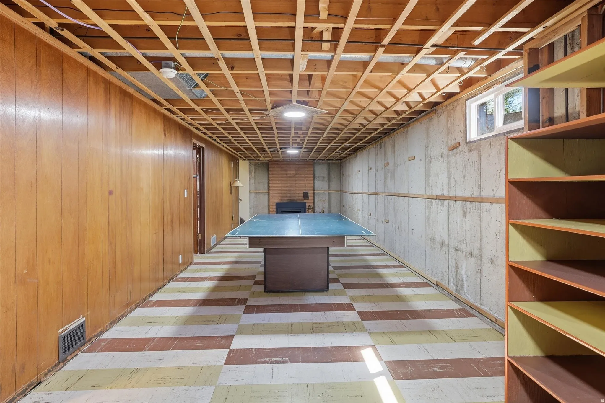 Spacious lower-level area featuring wood paneling, exposed ceiling joists, a checkered floor, and built-in shelving