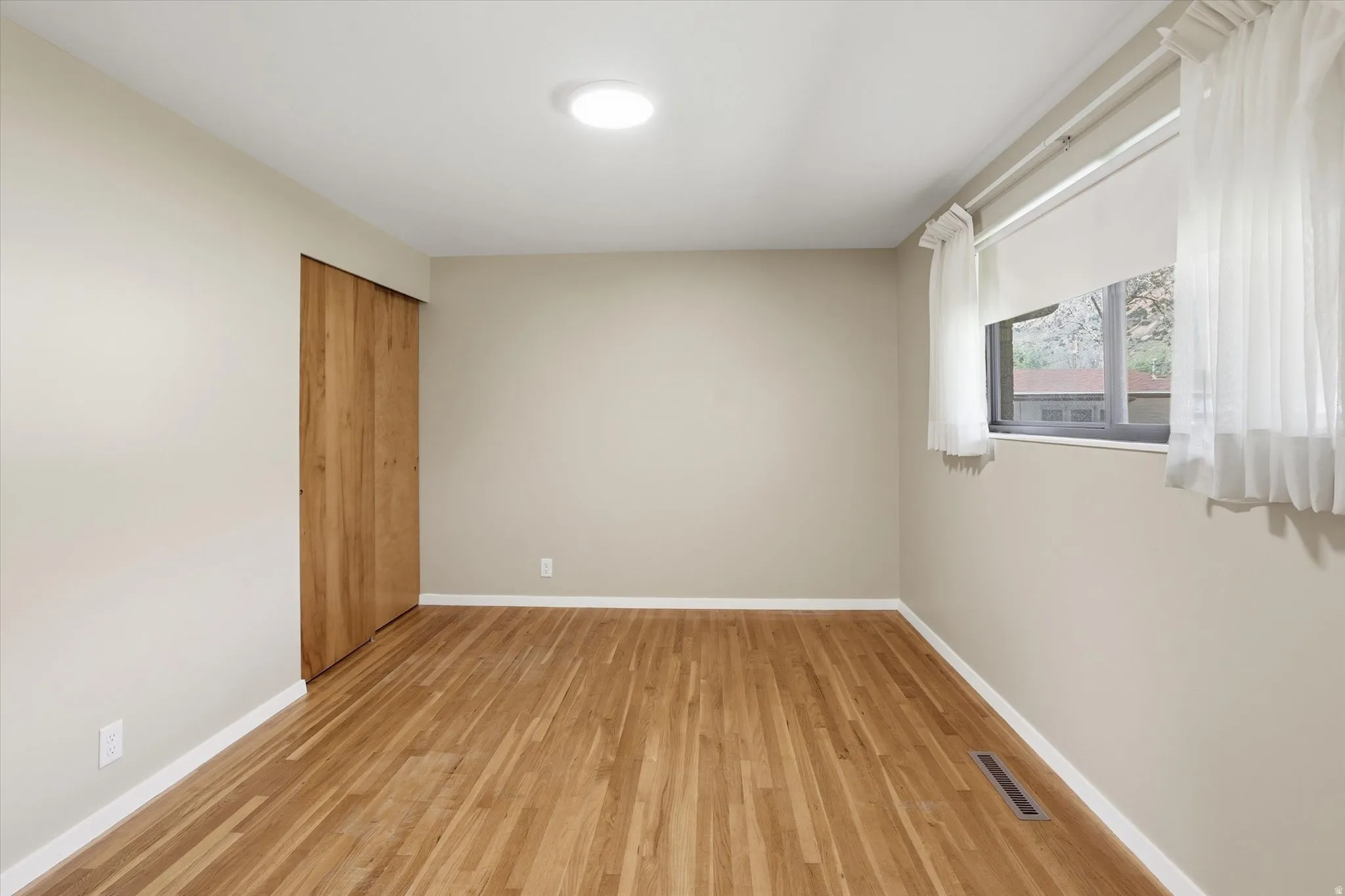 Room featuring wood-finish flooring, neutral wall paint, a window with roller shade and sheer curtains, a recessed ceiling light, and a bi-fold wood closet door