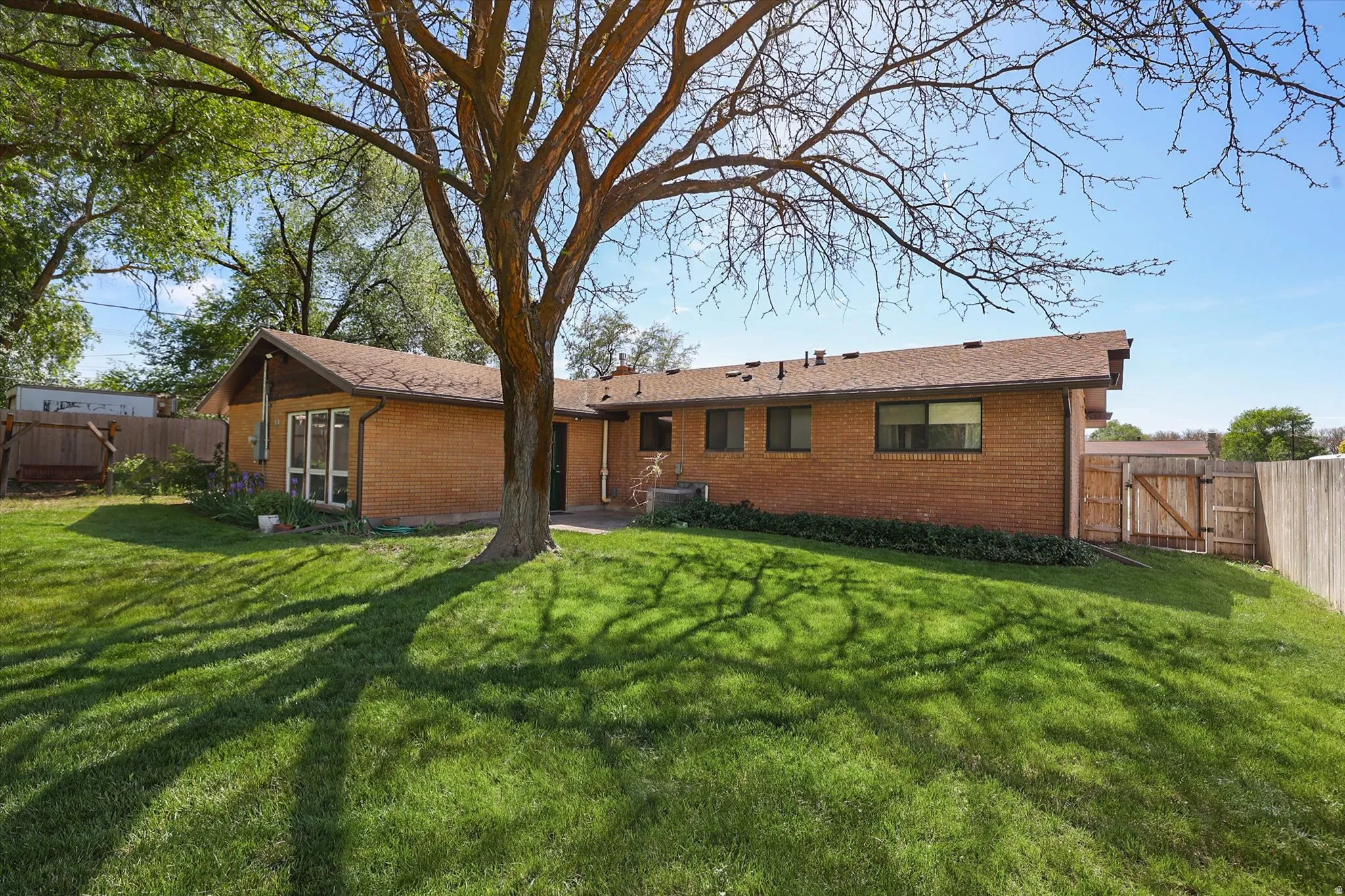 Expansive grassy backyard featuring a mature shade tree, a brick exterior, a brown shingle roof, and a wood privacy fence