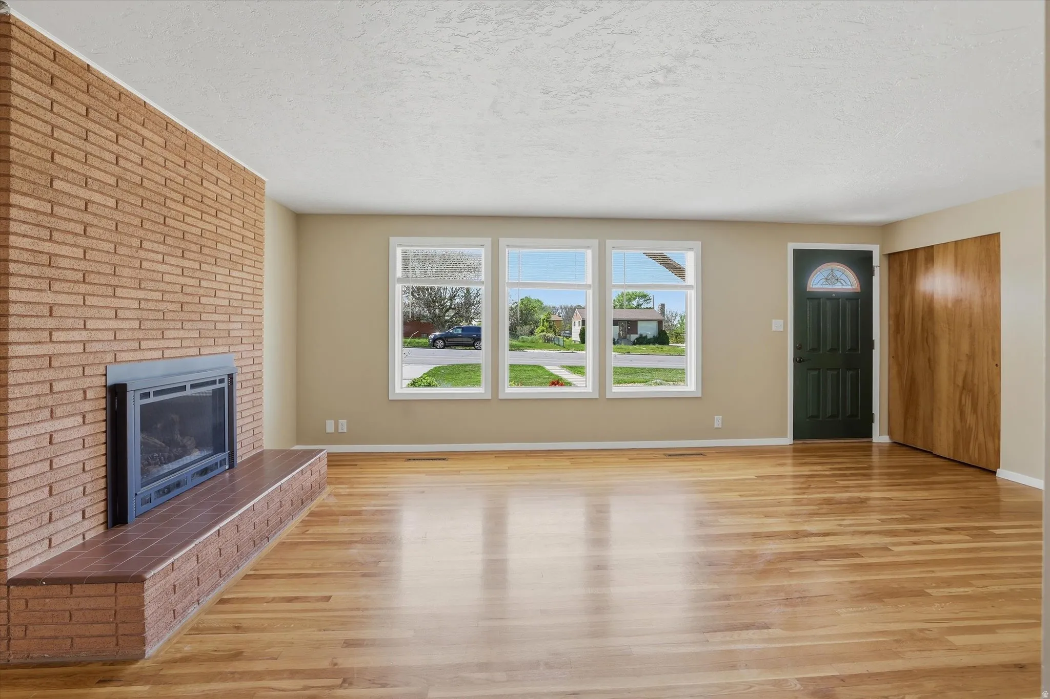 Spacious living area featuring polished wood-finish flooring, a brick-clad fireplace with hearth, multiple windows, a paneled entry door with an arched glass insert, and built-in closet storage with bi-fold wood doors