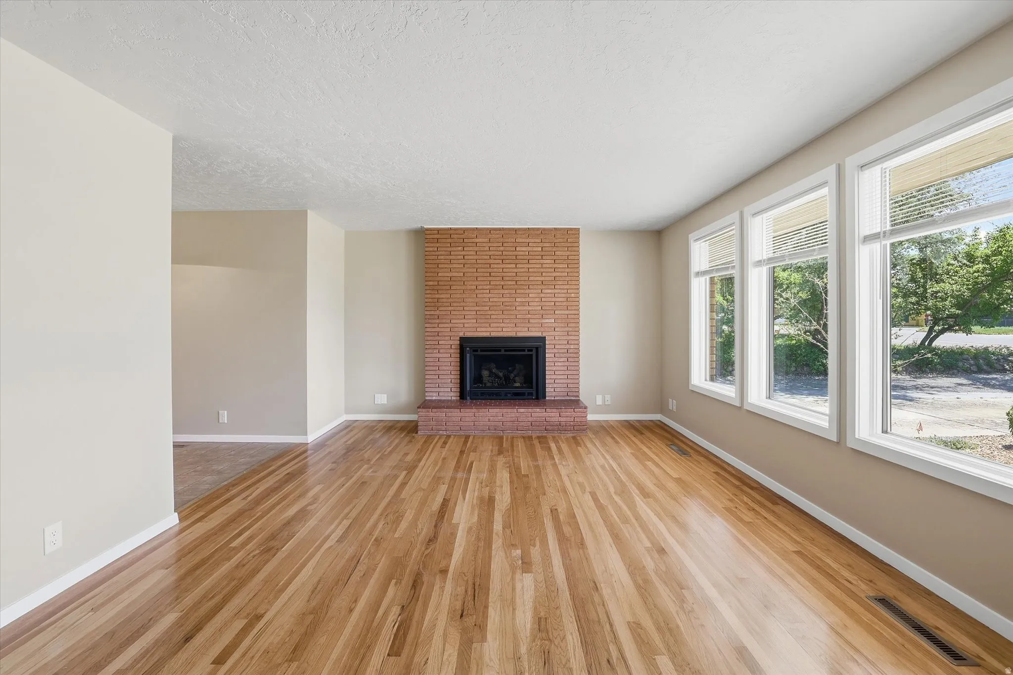 Spacious living area featuring light wood-finish flooring, a brick fireplace with a black insert, and three large windows providing abundant natural light