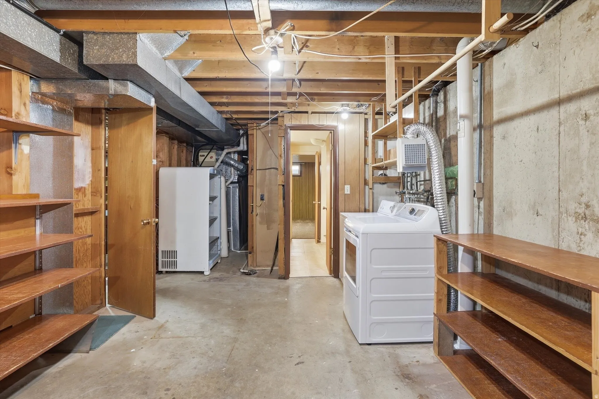 Unfinished basement featuring concrete flooring, exposed ceiling joists, built-in wooden shelving, and a laundry area with appliances