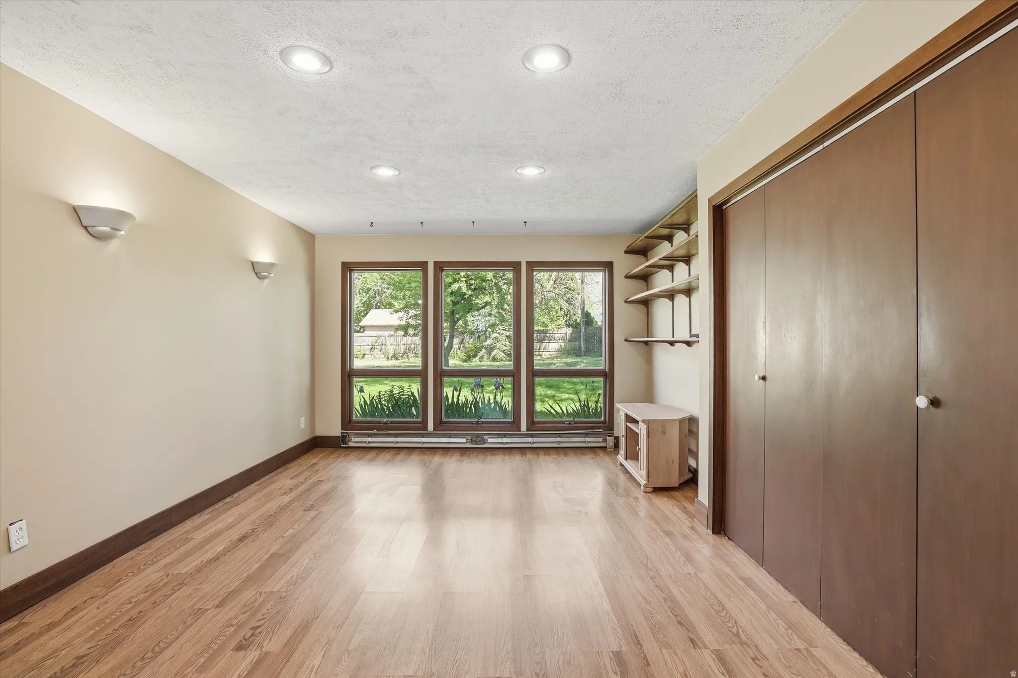 Light-filled room featuring three large windows, wood-finish flooring, recessed ceiling lights, and two built-in closets