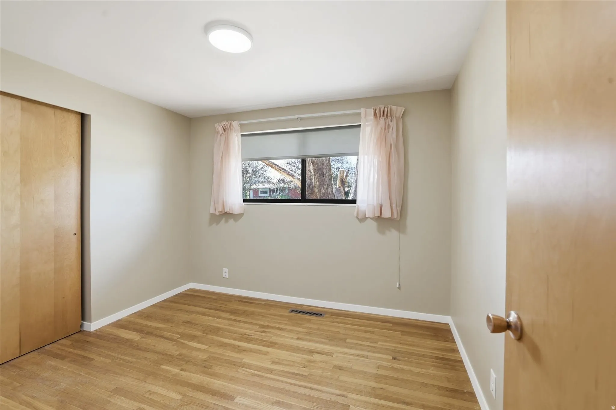 Room with wood-finish flooring, a window with a roller shade, and a flush-mount ceiling light