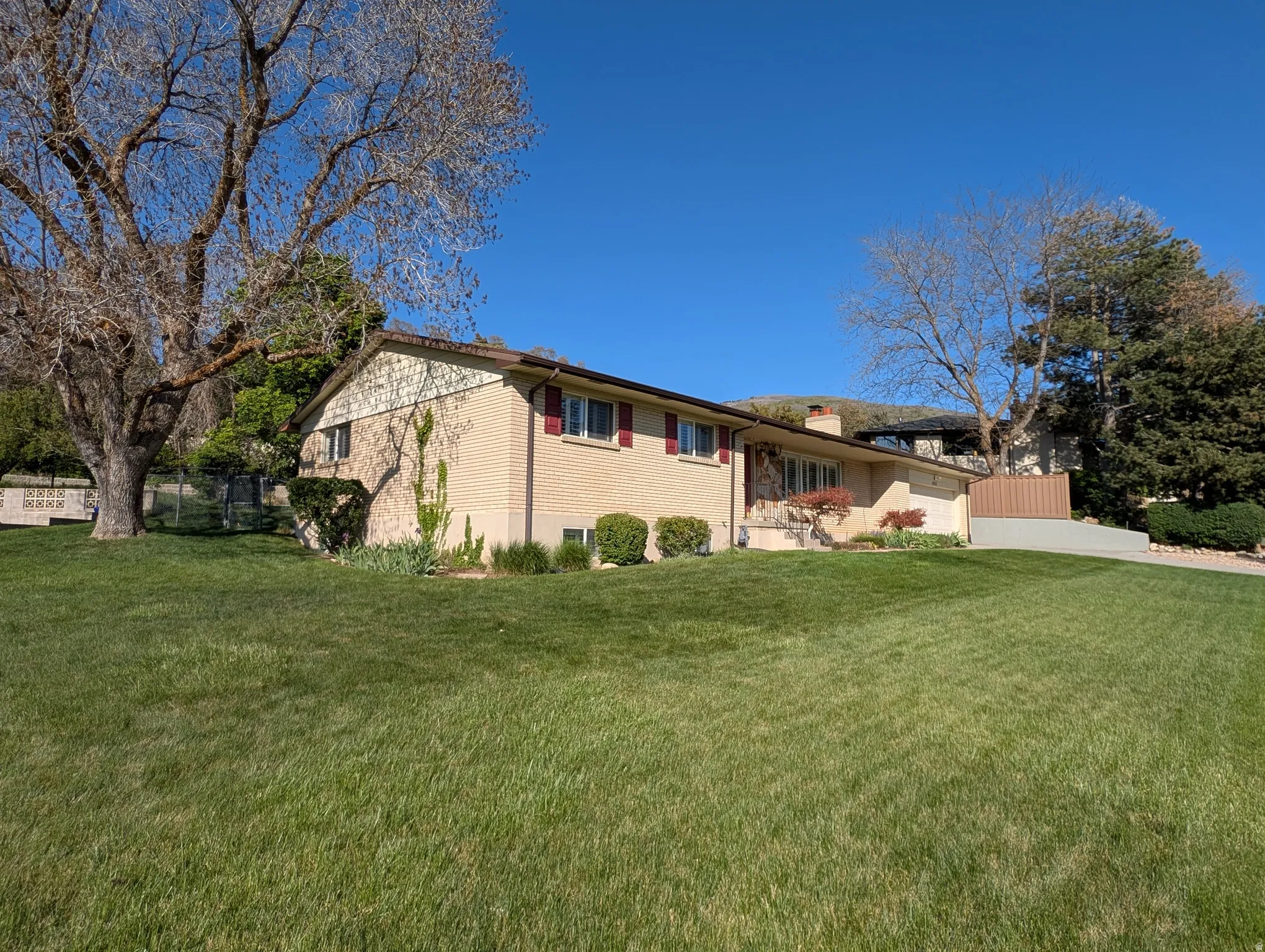 Brick exterior with light-colored siding, a sloped front lawn, mature trees, and a concrete driveway