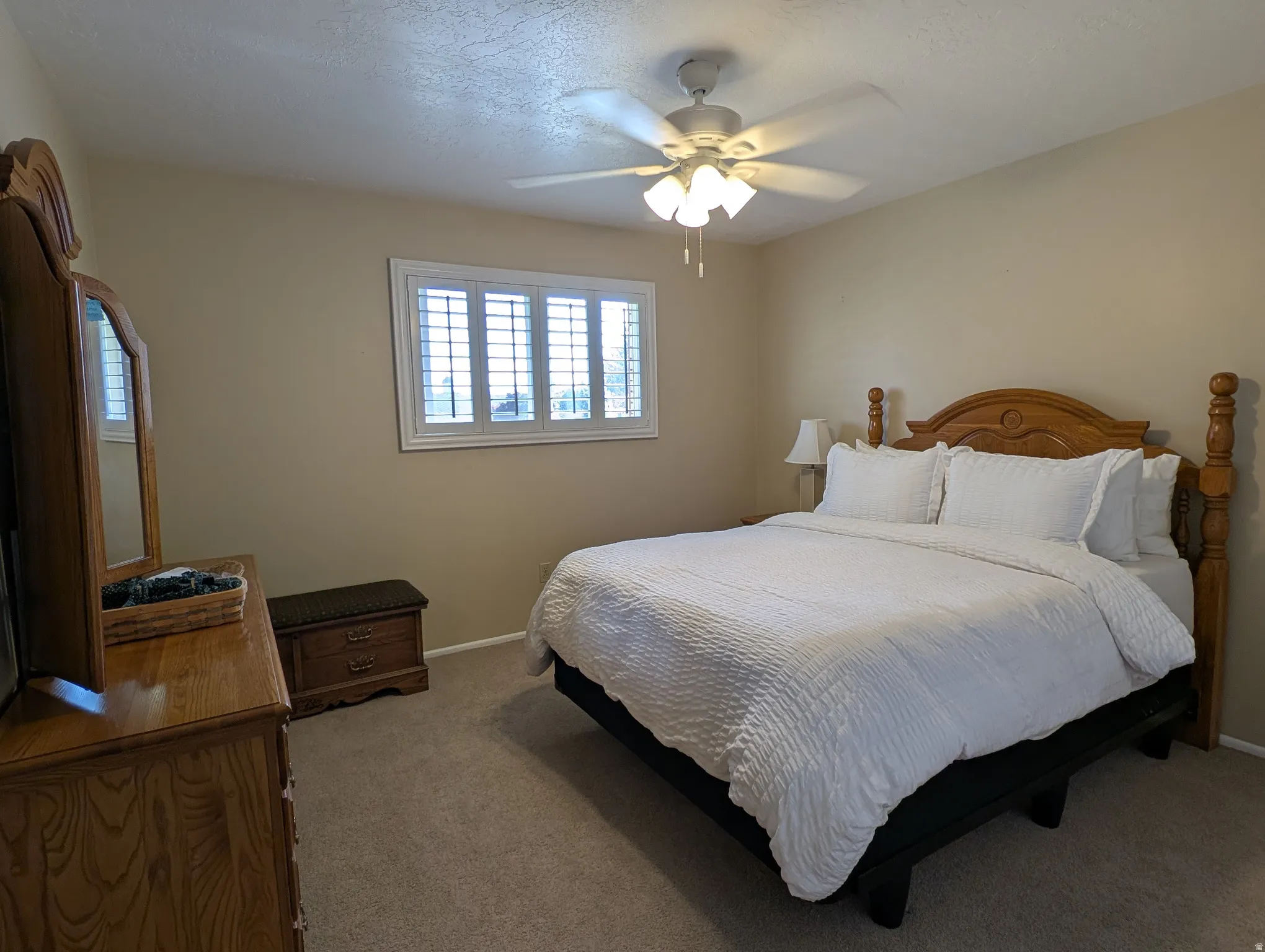 Carpeted bedroom featuring a ceiling fan with light fixture, window with white plantation shutters, and light beige wall paint