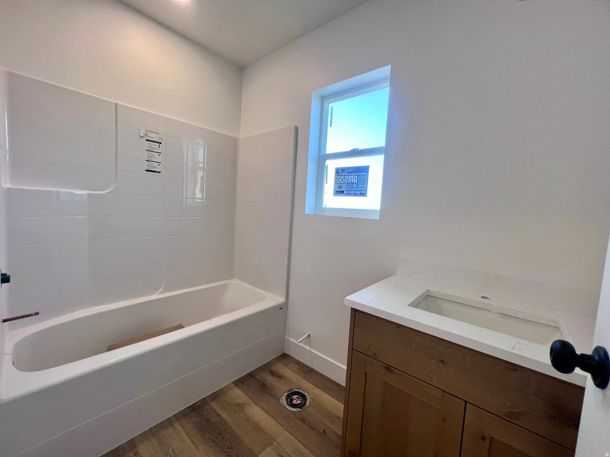 Bathroom featuring a white bathtub with tile surround, wood-finish flooring, a wood vanity with a white countertop and integrated sink, and a window providing natural light
