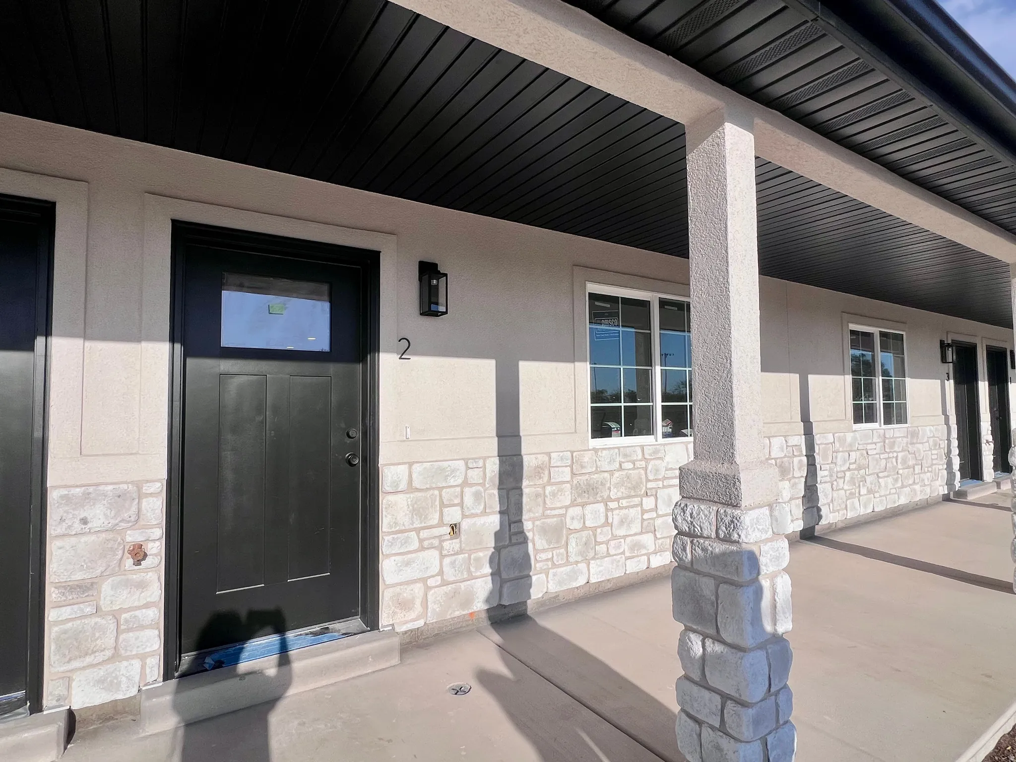 Exterior featuring a covered porch with textured columns, black entry door with transom window, stone facade accents, stucco siding, and white-framed windows