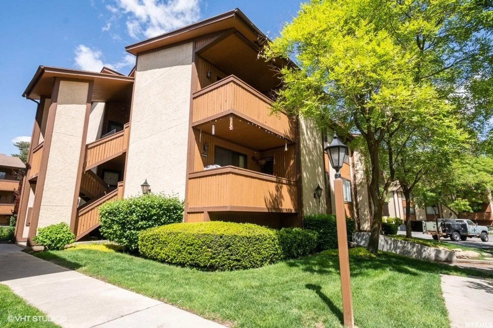 Multi-story building featuring stucco siding, individual balconies with wood railings, exterior staircases, and mature landscaping