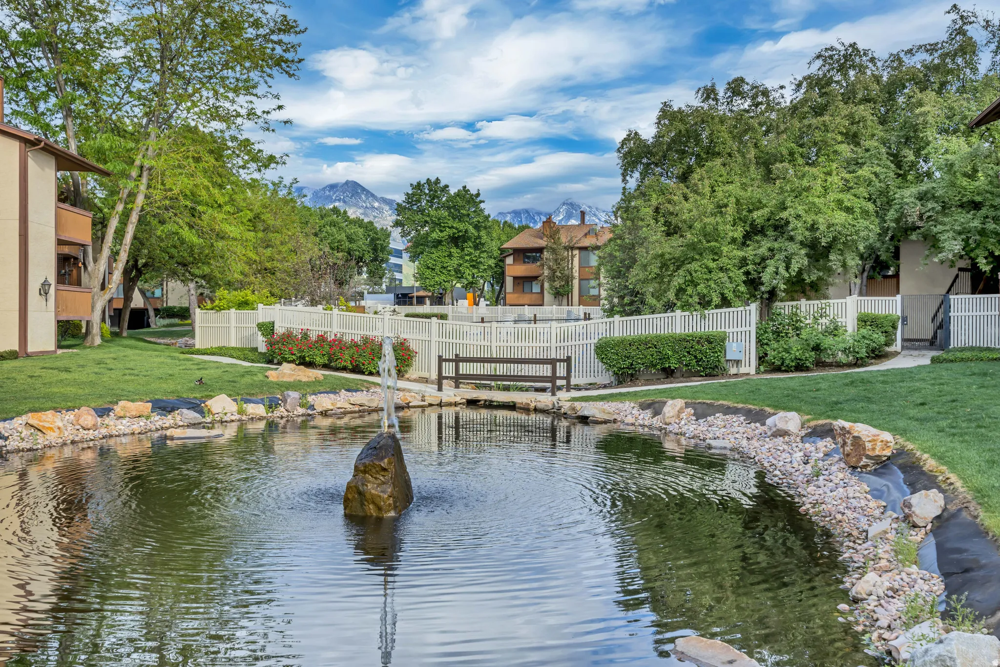 Serene water feature with a rock fountain centerpiece, surrounded by manicured green spaces and a white picket fence