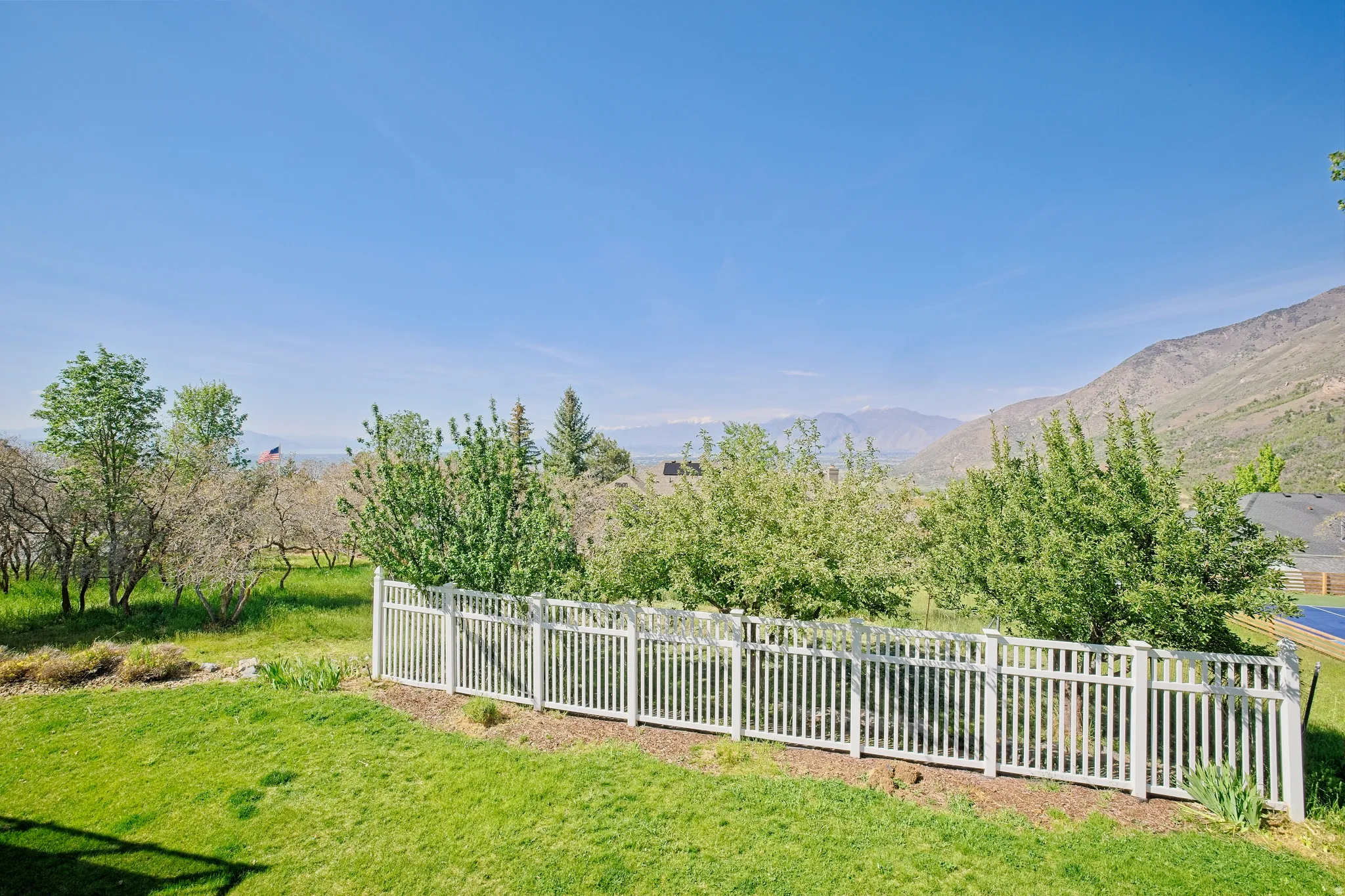 Expansive green lawn with a white picket fence bordering mature trees, set against a backdrop of distant mountains