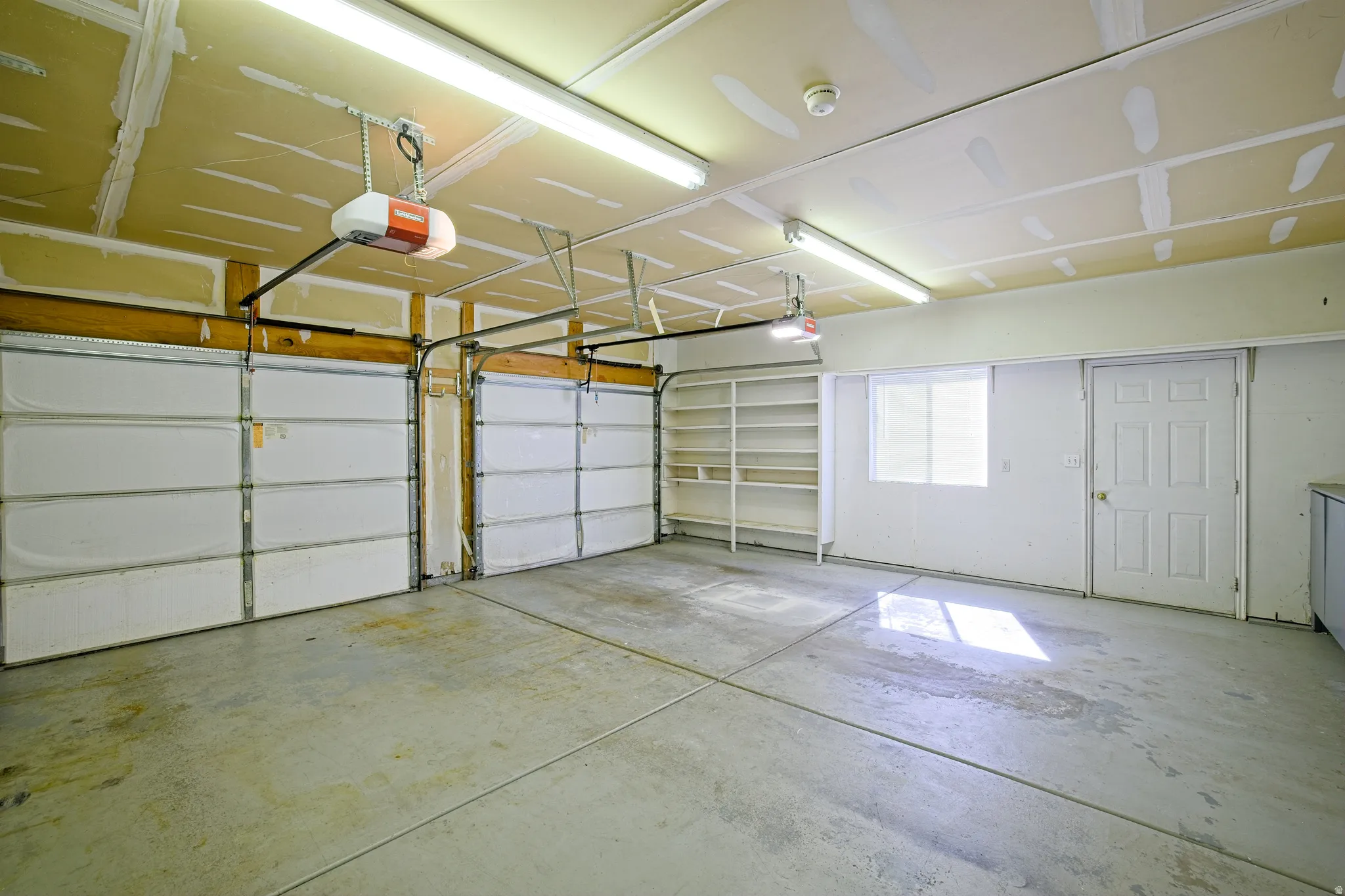 Spacious garage featuring a concrete floor, two individual garage doors with automatic openers, fluorescent lighting, built-in shelving, and a side entry door