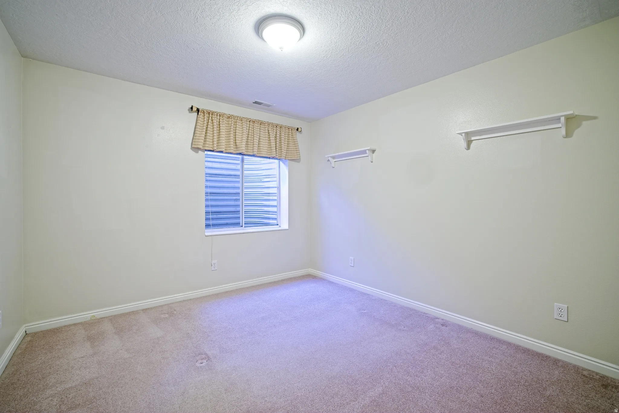 Carpeted room featuring a single window with blinds, ceiling-mounted light fixture, and two wall-mounted shelves