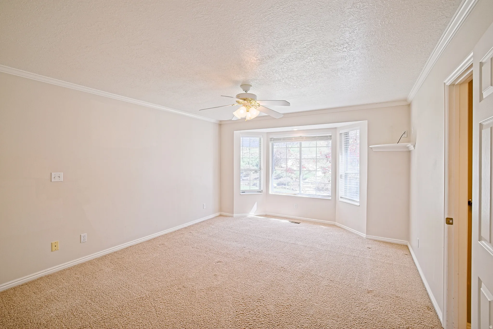 Carpeted room featuring a bay window with blinds, ceiling fan, crown molding, and a built-in wall shelf