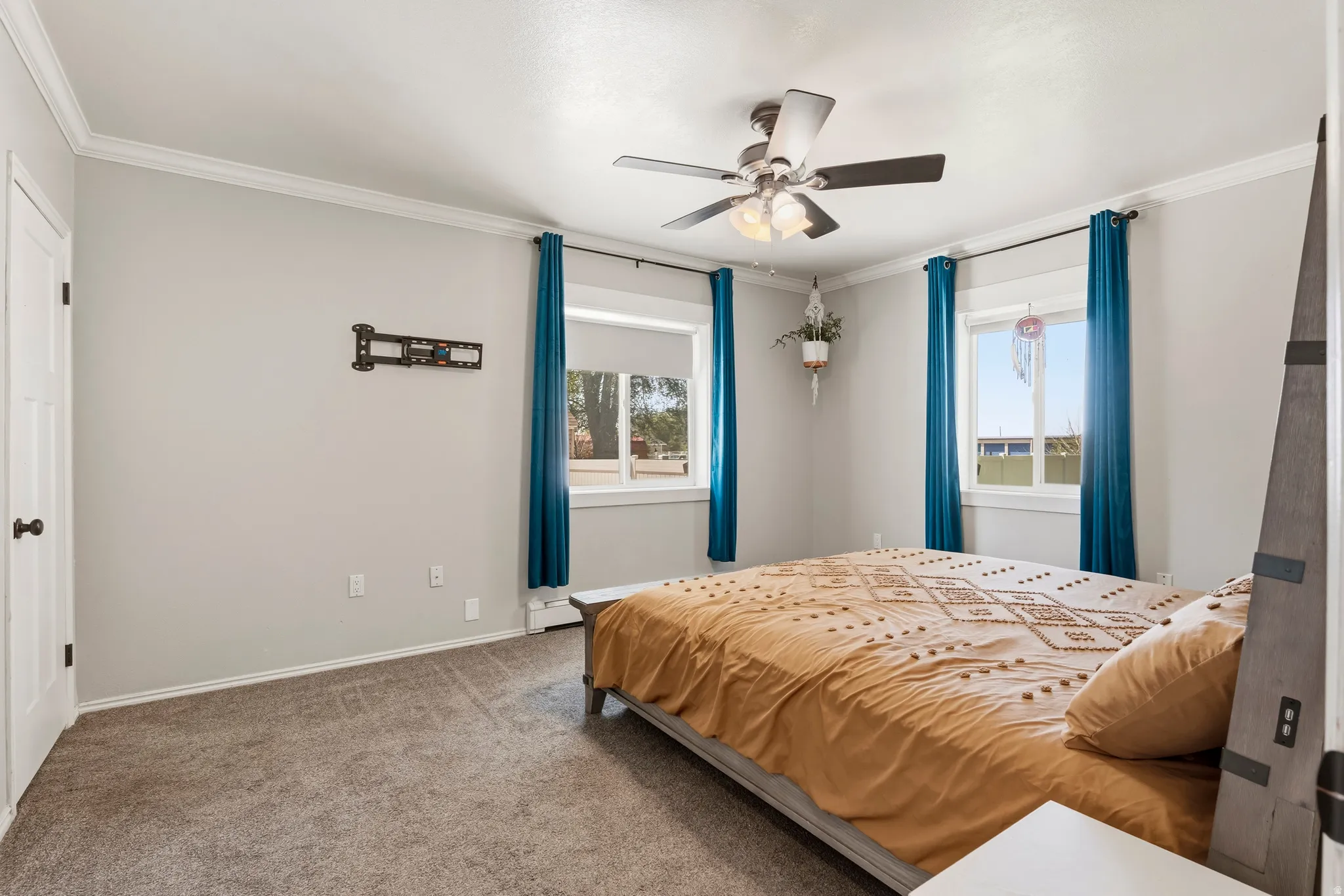 Carpeted bedroom featuring two windows with white trim, crown molding, a ceiling fan with integrated lighting, and a wall-mounted TV bracket