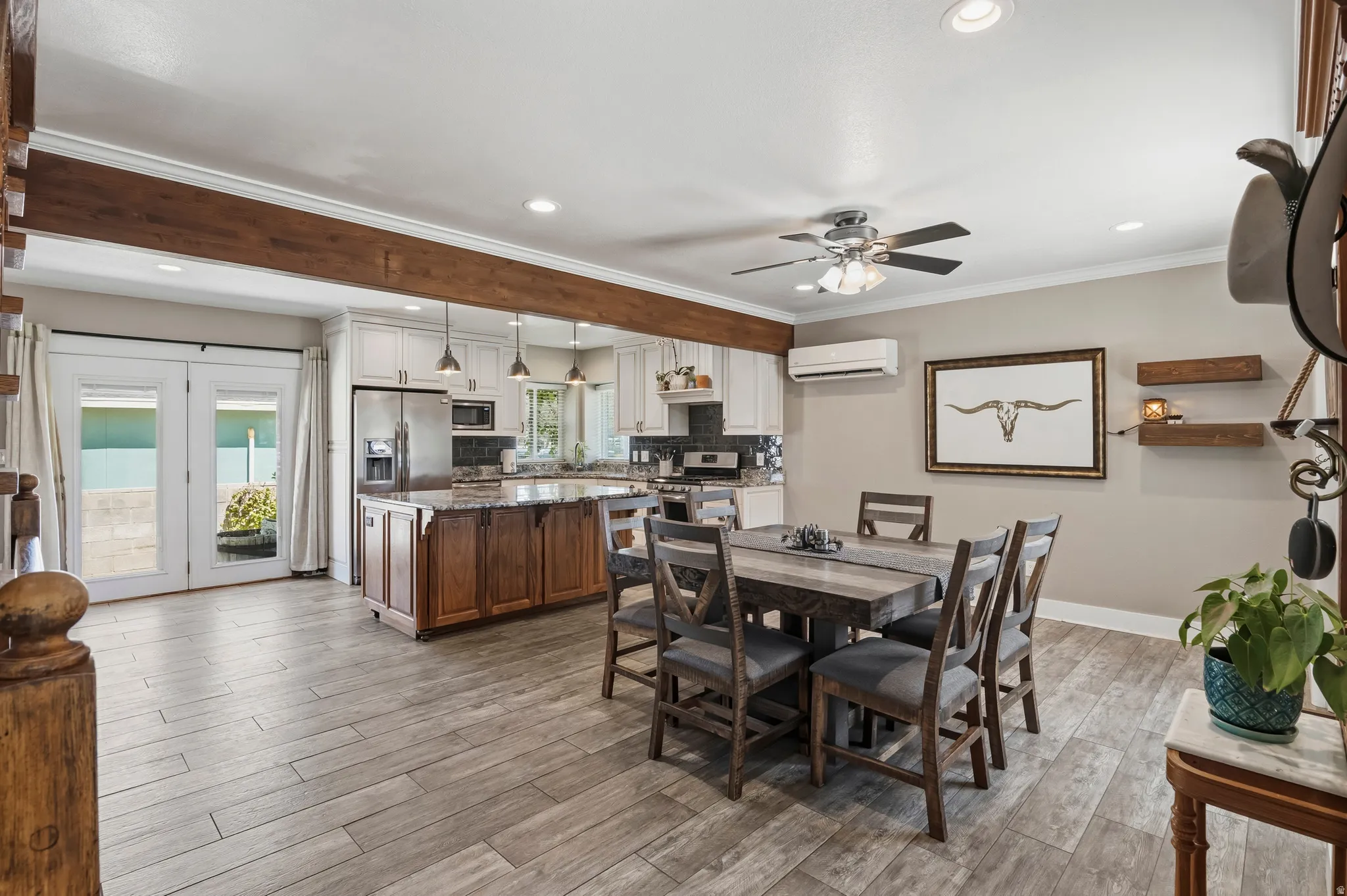 Open-concept kitchen and dining area featuring wood-finish flooring, a wood-beam ceiling accent, a central island with a stone countertop, white cabinetry with pendant lighting, and double French doors opening to an exterior paved area